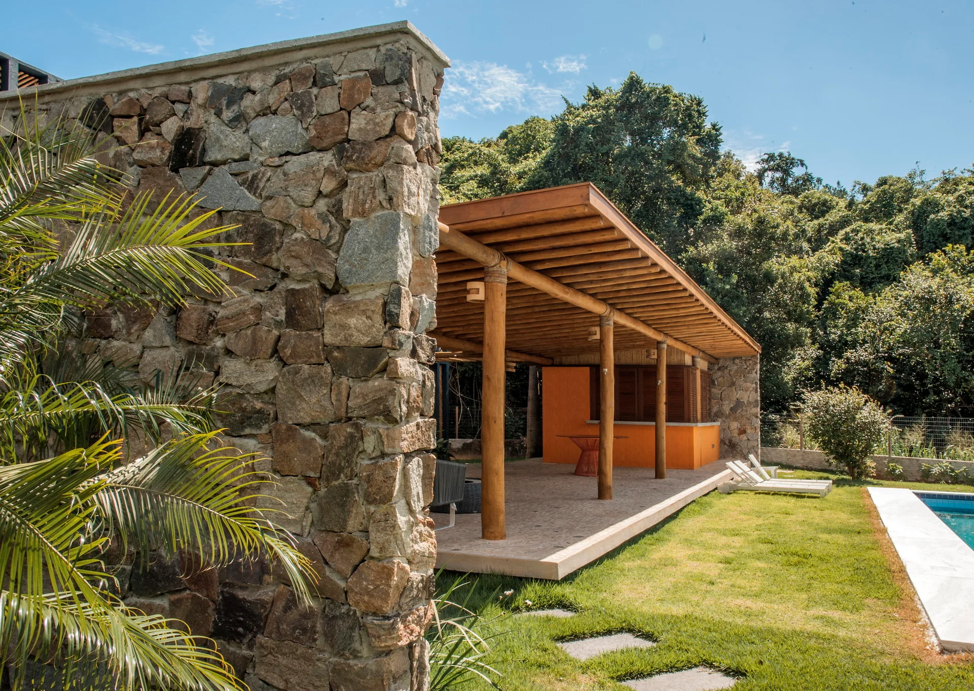 A covered outdoor patio featuring a rustic stone wall, a wooden timber canopy, and an orange accent counter next to a grassy lawn and swimming pool.