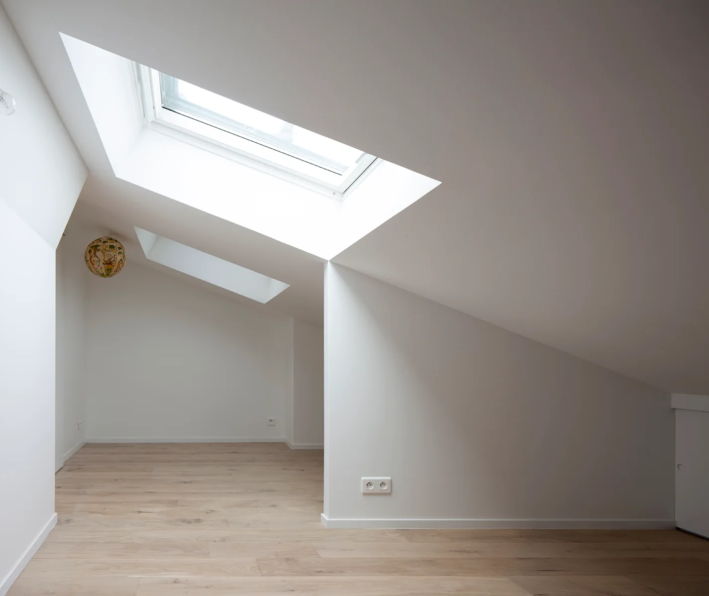 An empty attic space features light wood plank flooring, sloped white walls, and two rectangular skylights filtering in natural light.