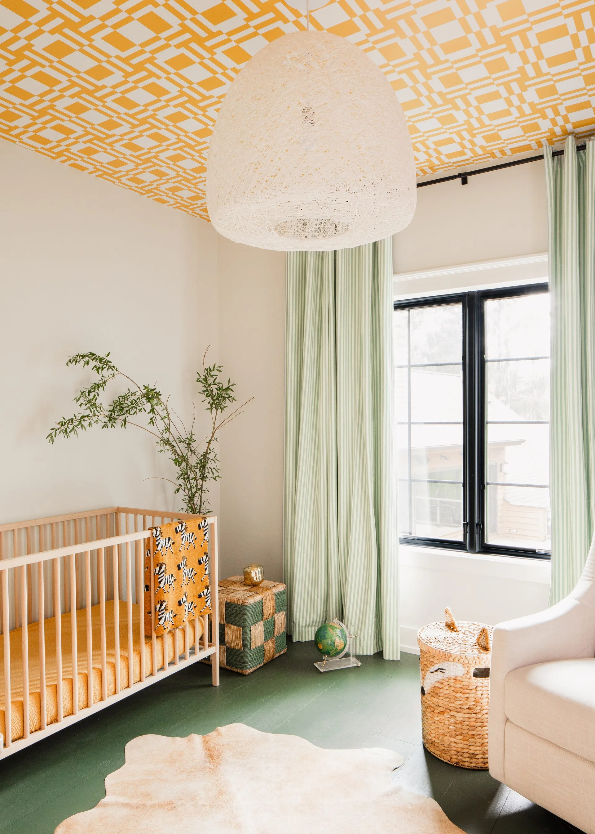 A light-filled nursery features a wooden crib, striped green curtains, a yellow patterned ceiling, and a dark green painted wood floor.