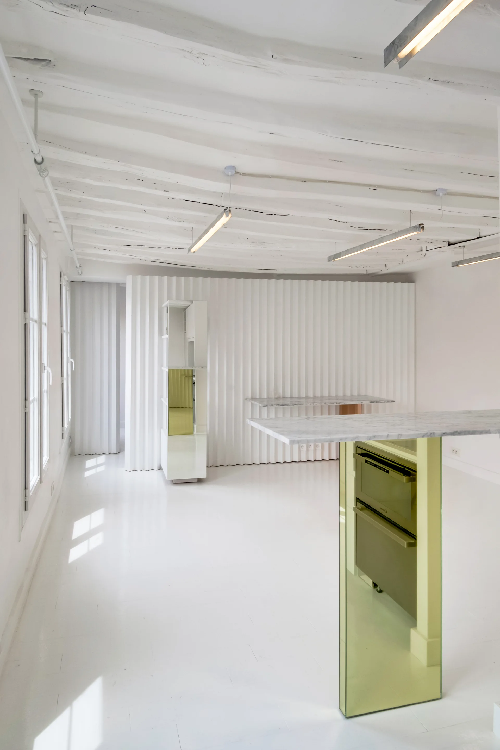 A minimalist white kitchen featuring a marble countertop island with a yellow glass base, white beamed ceiling, and a wavy corrugated accent wall.