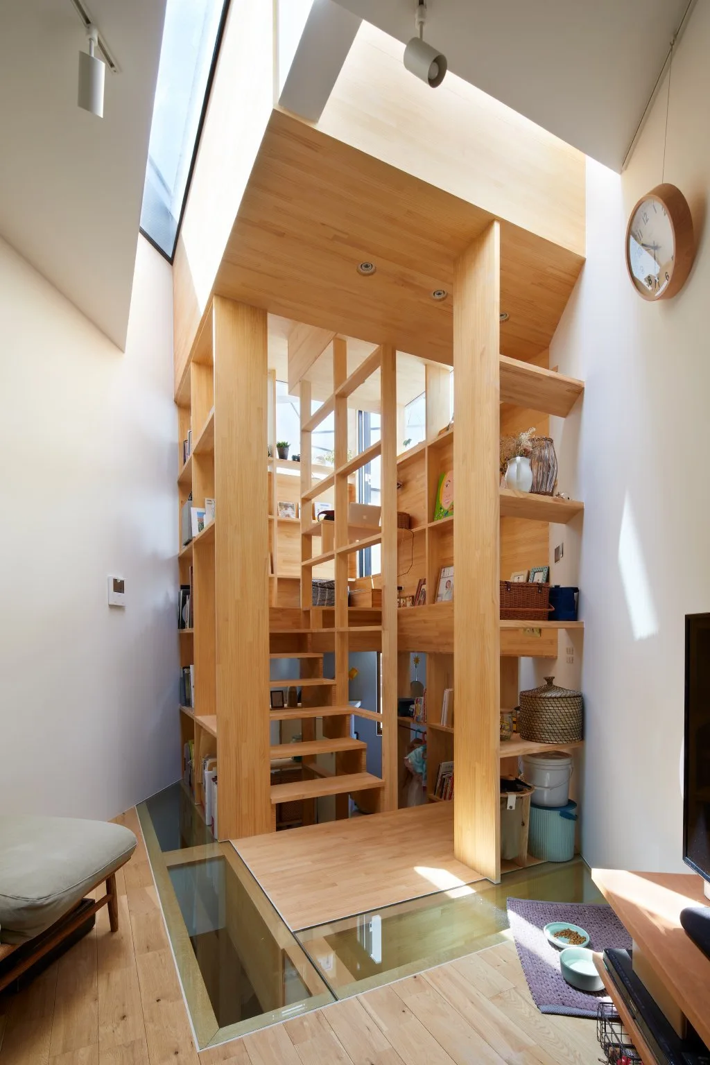A light wood tower structure combining stairs and open shelves stands over a glass floor panel next to white walls.