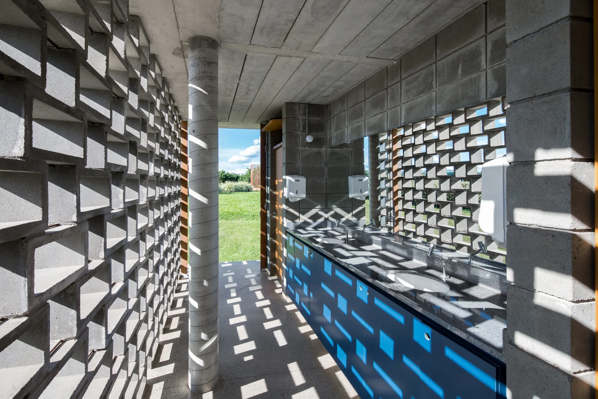 A modern concrete restroom features a breeze block wall casting geometric shadow patterns over a long blue vanity.