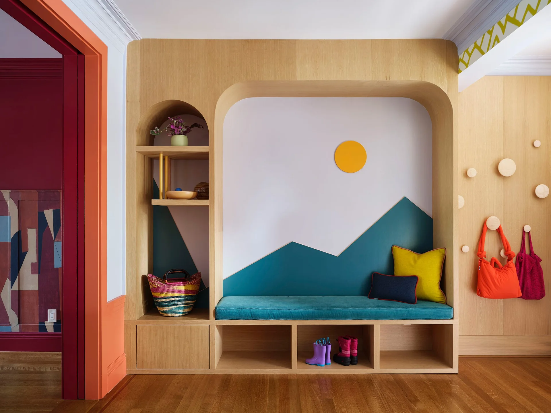 A light wood built-in mudroom bench features integrated storage cubbies, a playful painted mountain mural, and matching wall peg hooks.