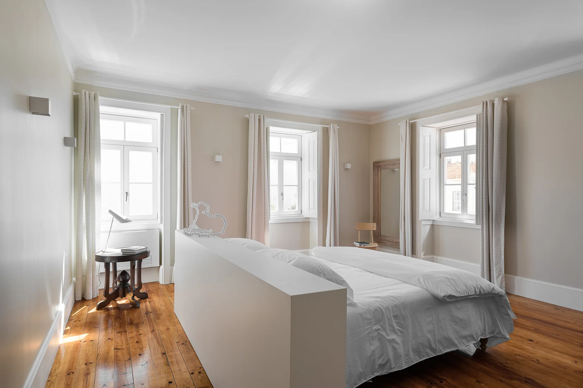 A minimalist bedroom featuring a central white partition wall behind a bed, warm wood floors, and beige walls with three draped windows.