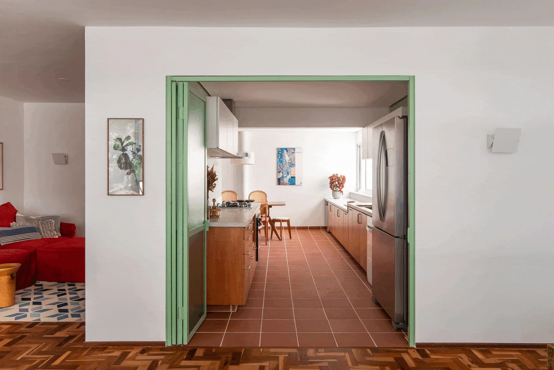 A view through a mint green metal door frame into a kitchen featuring warm wood cabinetry, terracotta tile flooring, and a stainless steel refrigerator.