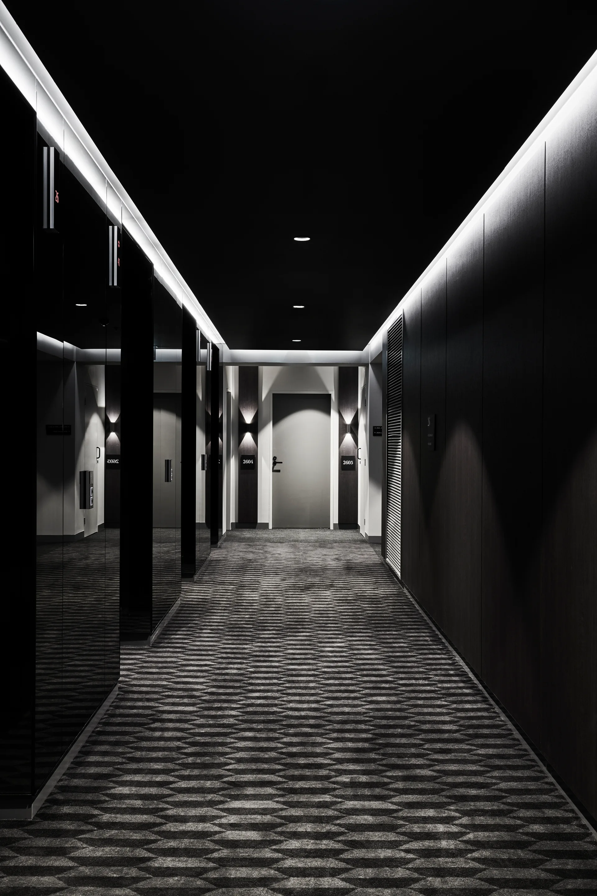A long dark hallway with black walls, a gray geometric diamond patterned carpet, and bright cove lighting lines on the ceiling.