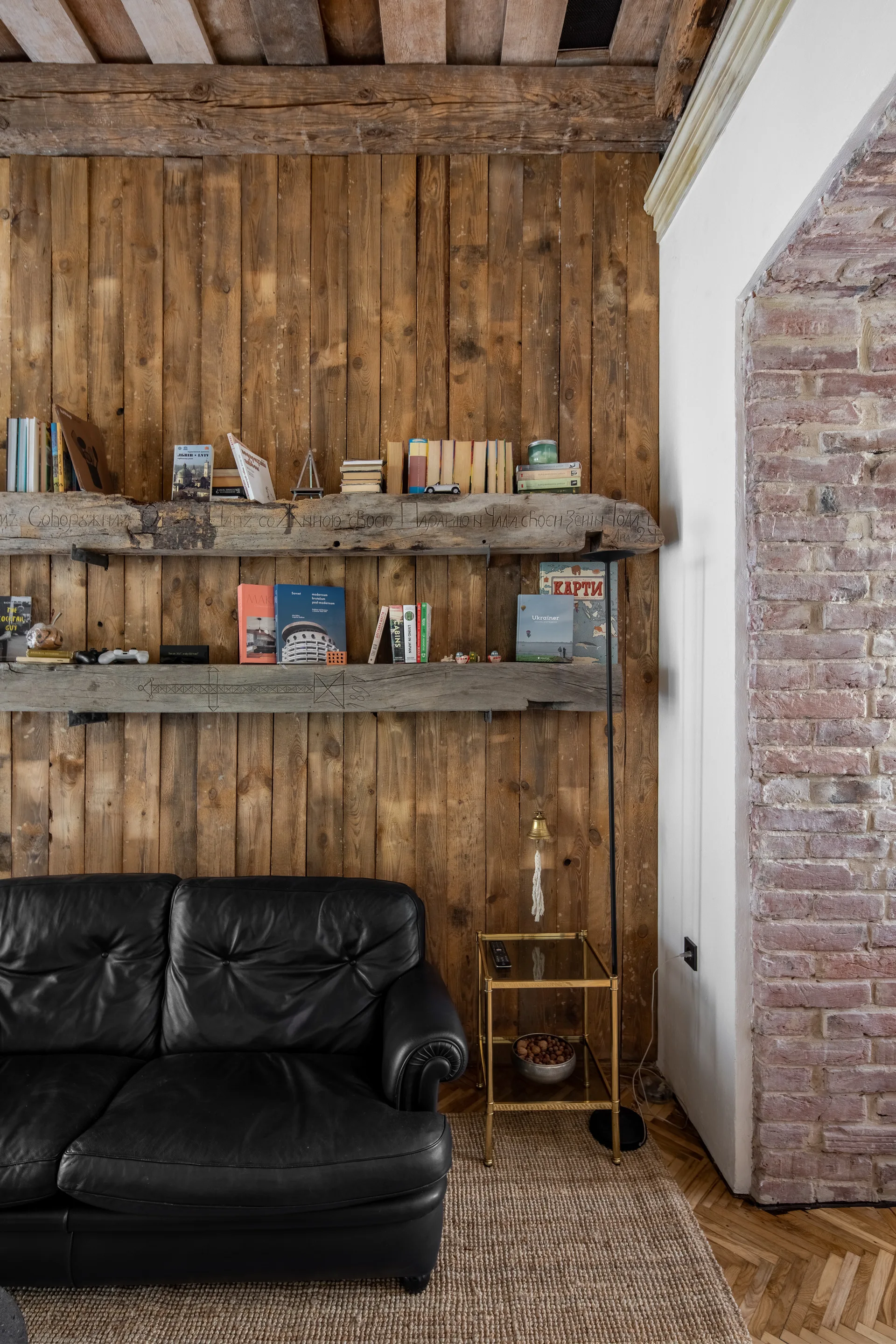 A black leather sofa sits against a vertical wood paneled wall featuring two thick, raw timber floating shelves filled with books.
