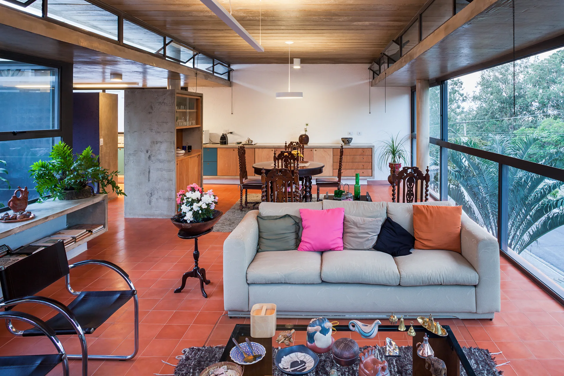 An open living room featuring a cream sofa with bright pillows, a wooden ceiling, terracotta tile floors, and large panoramic windows.