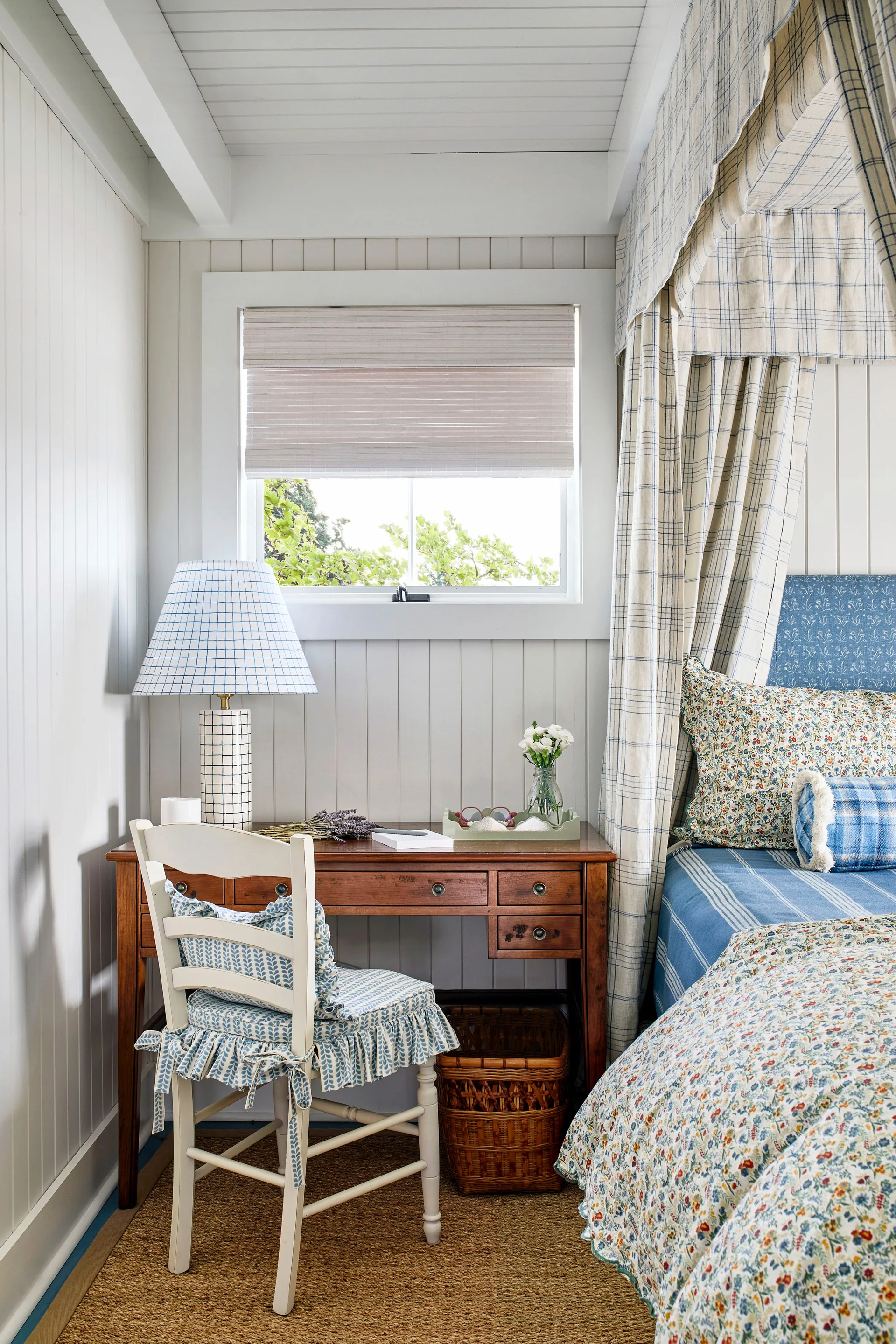 A cottage bedroom features a canopy bed with blue and white patterned linens next to a wooden writing desk with a grid-pattern lamp and a white chair.