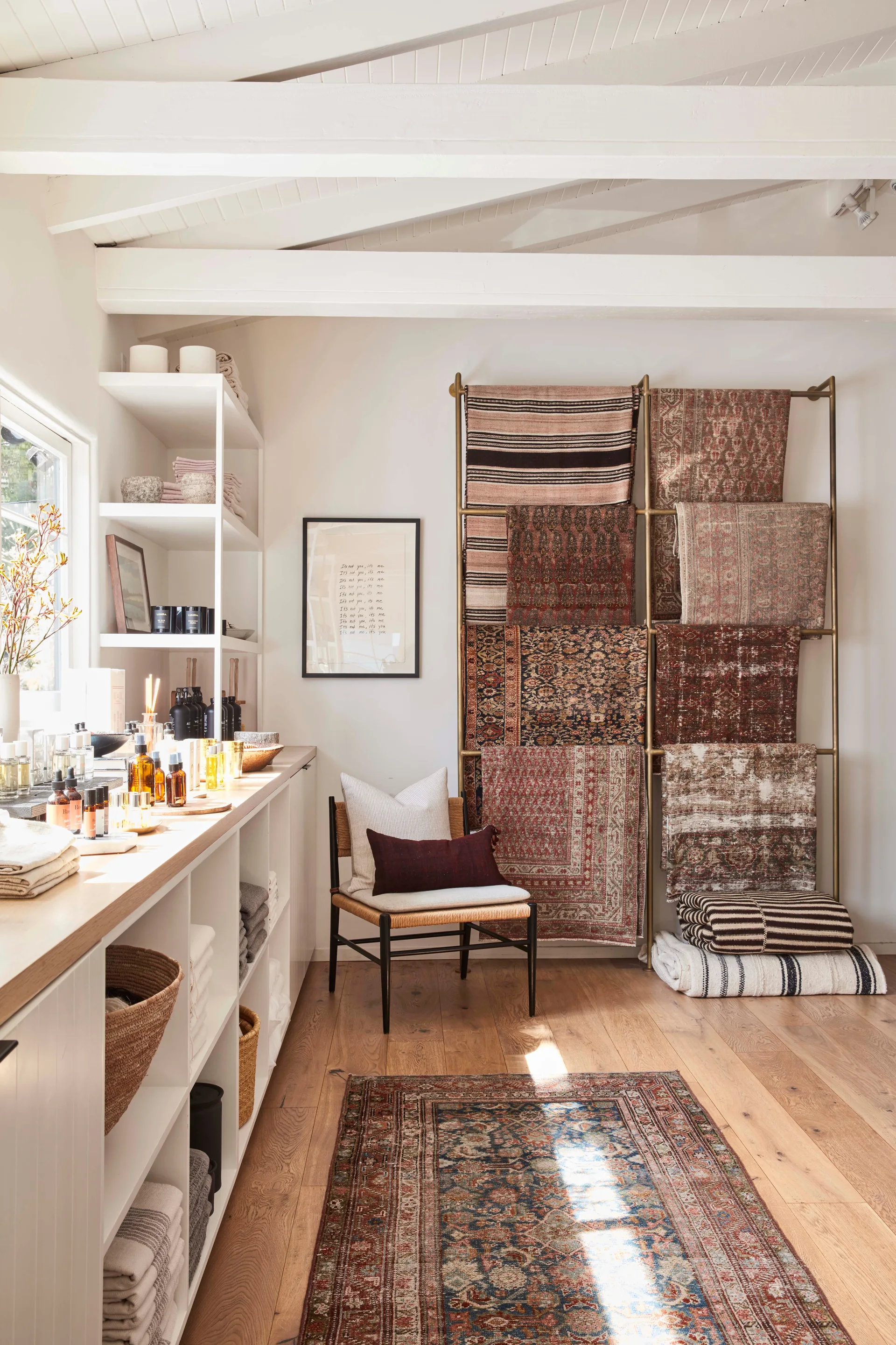 A well-lit boutique interior features a vertical brass rug display rack, white open shelving filled with home goods, and a rattan-back chair.