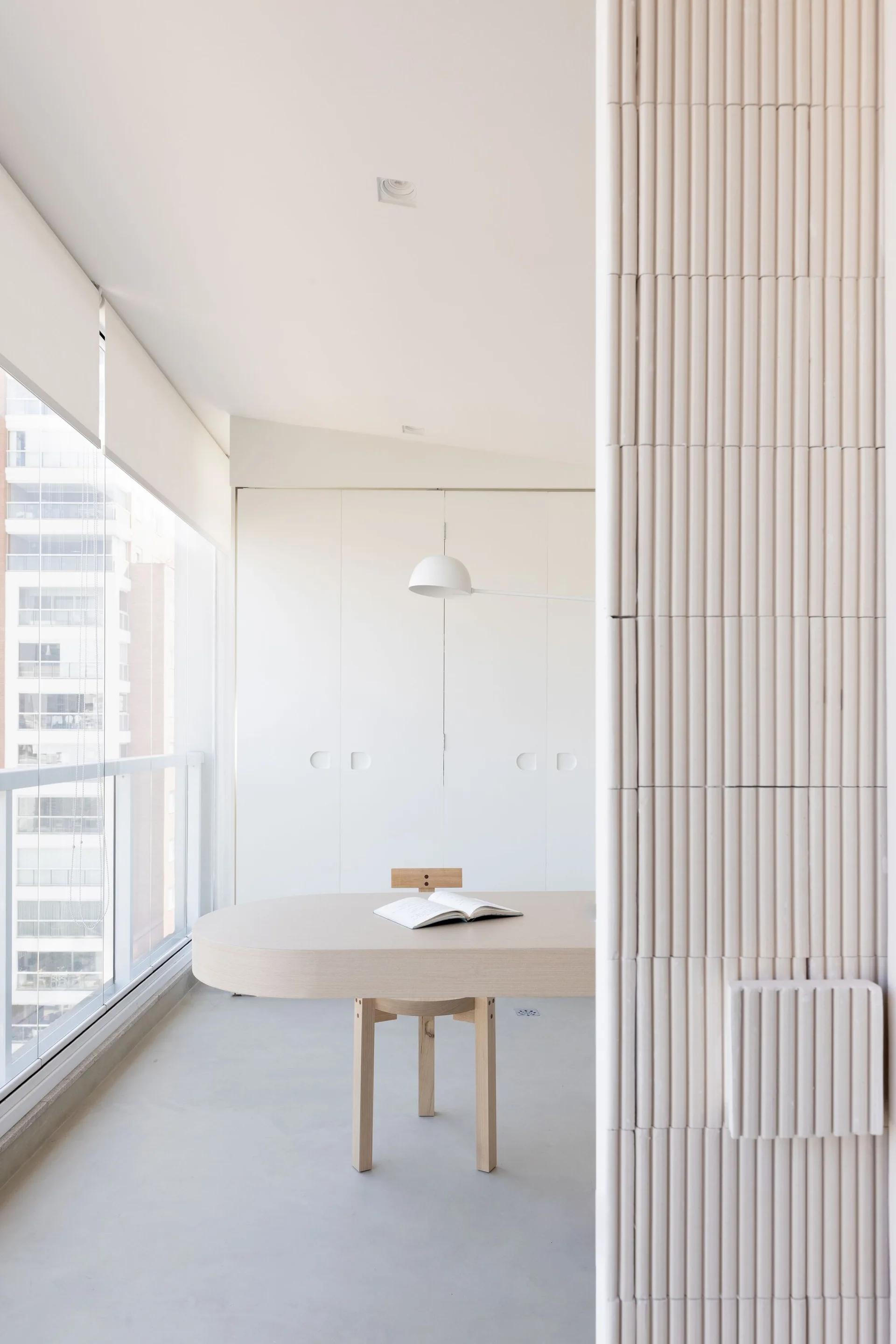 A minimalist dining space features an oval light wood table and chair next to a large window, with a textured fluted column in the foreground.