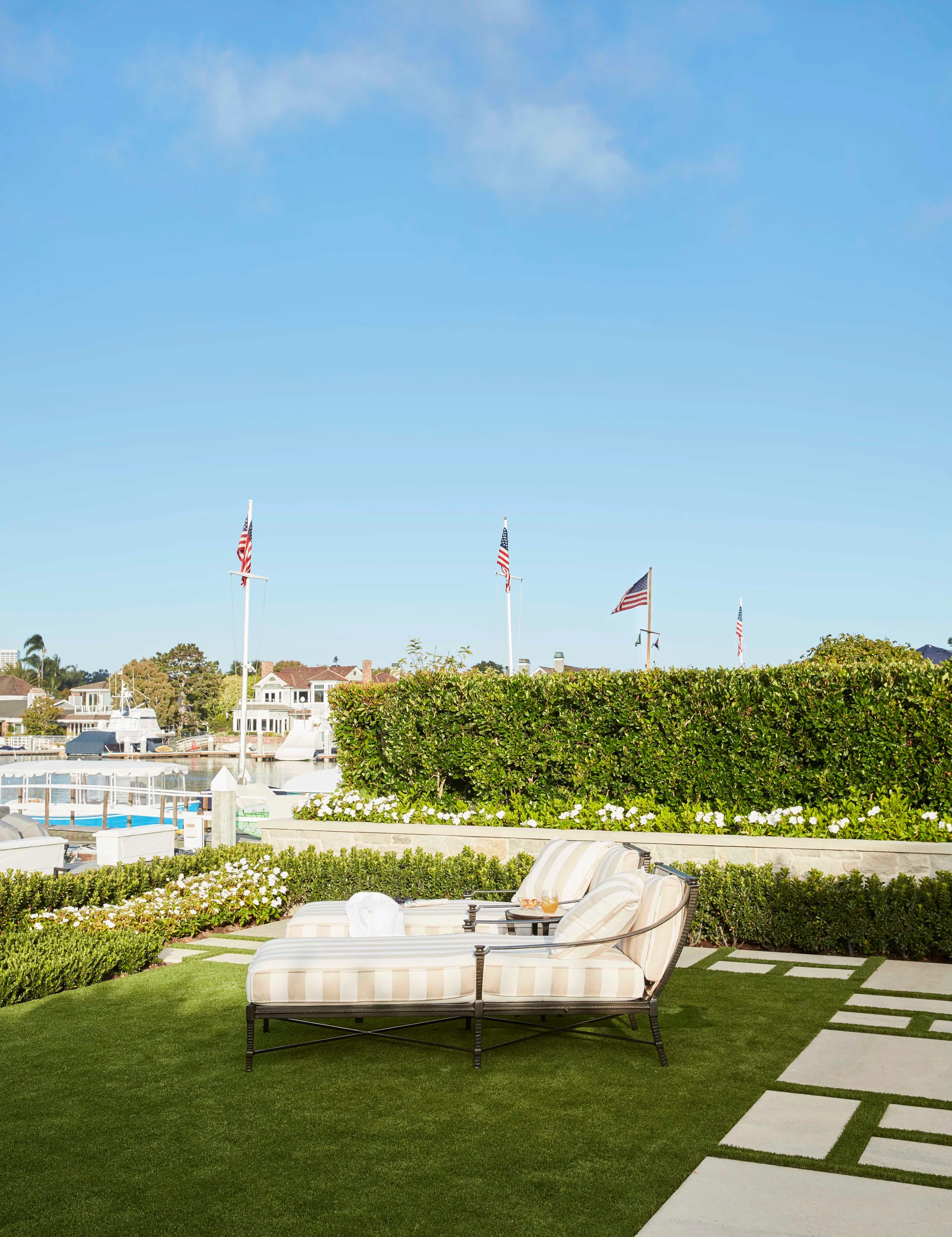 Two beige striped chaise lounges rest on a green lawn with stone pavers next to a waterfront hedge.