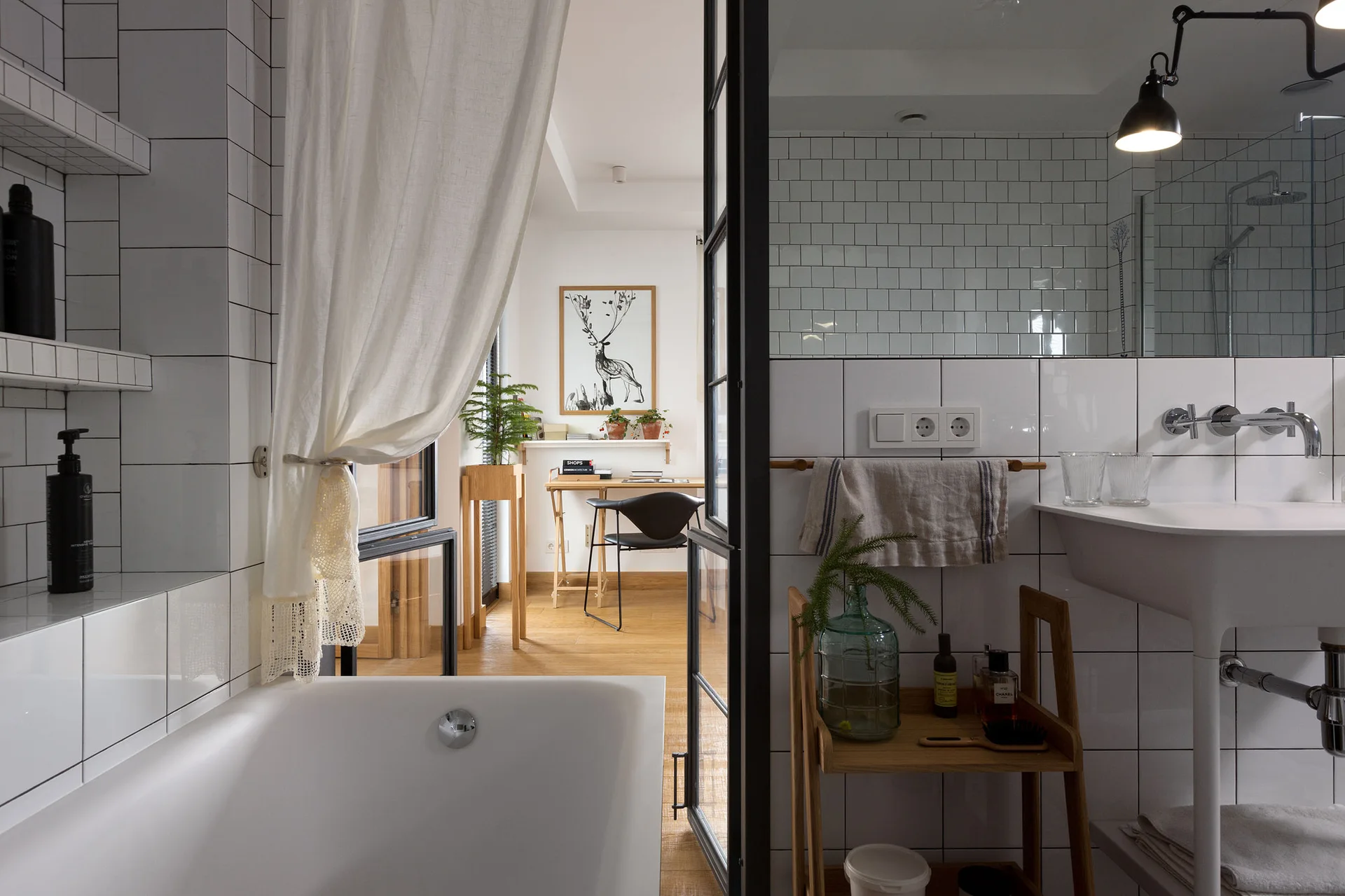 A modern bathroom with glossy white grid tiles and a black metal glass door opening to a light wood home office.