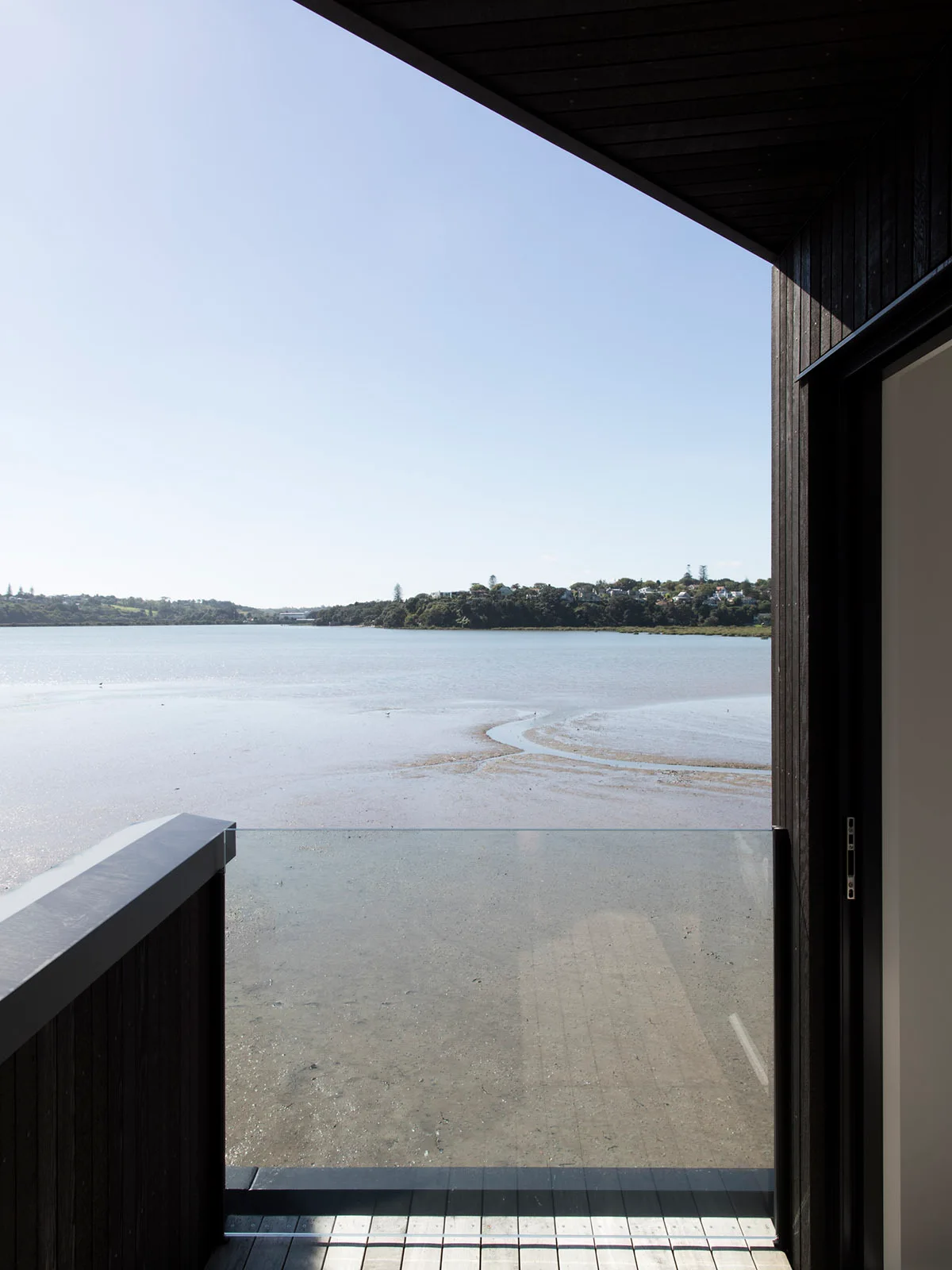 A modern balcony with dark vertical wood siding and a clear glass railing overlooks a wide tidal river under a bright blue sky.