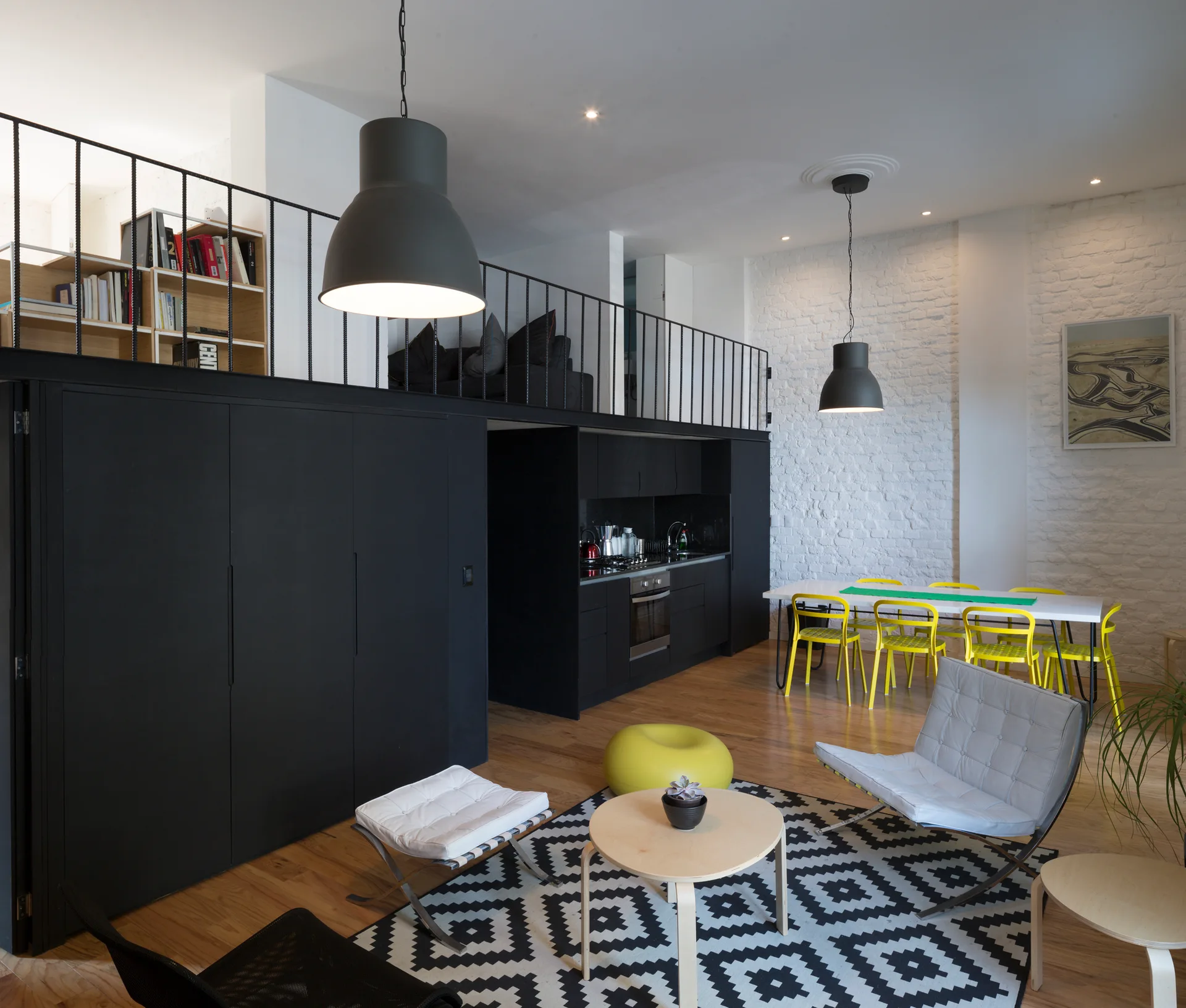 A modern loft apartment featuring a black storage structure under a mezzanine level, a white brick wall, and a dining table with yellow chairs.