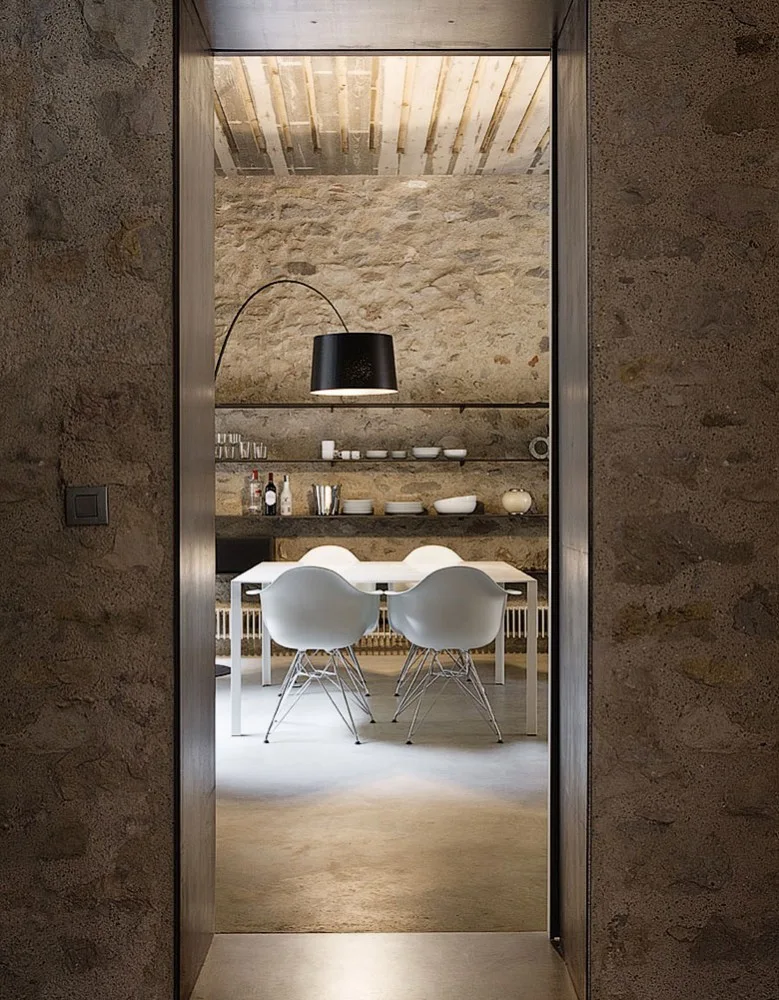A view through a doorway into a dining room featuring a white table with modern molded chairs against a textured stone wall.