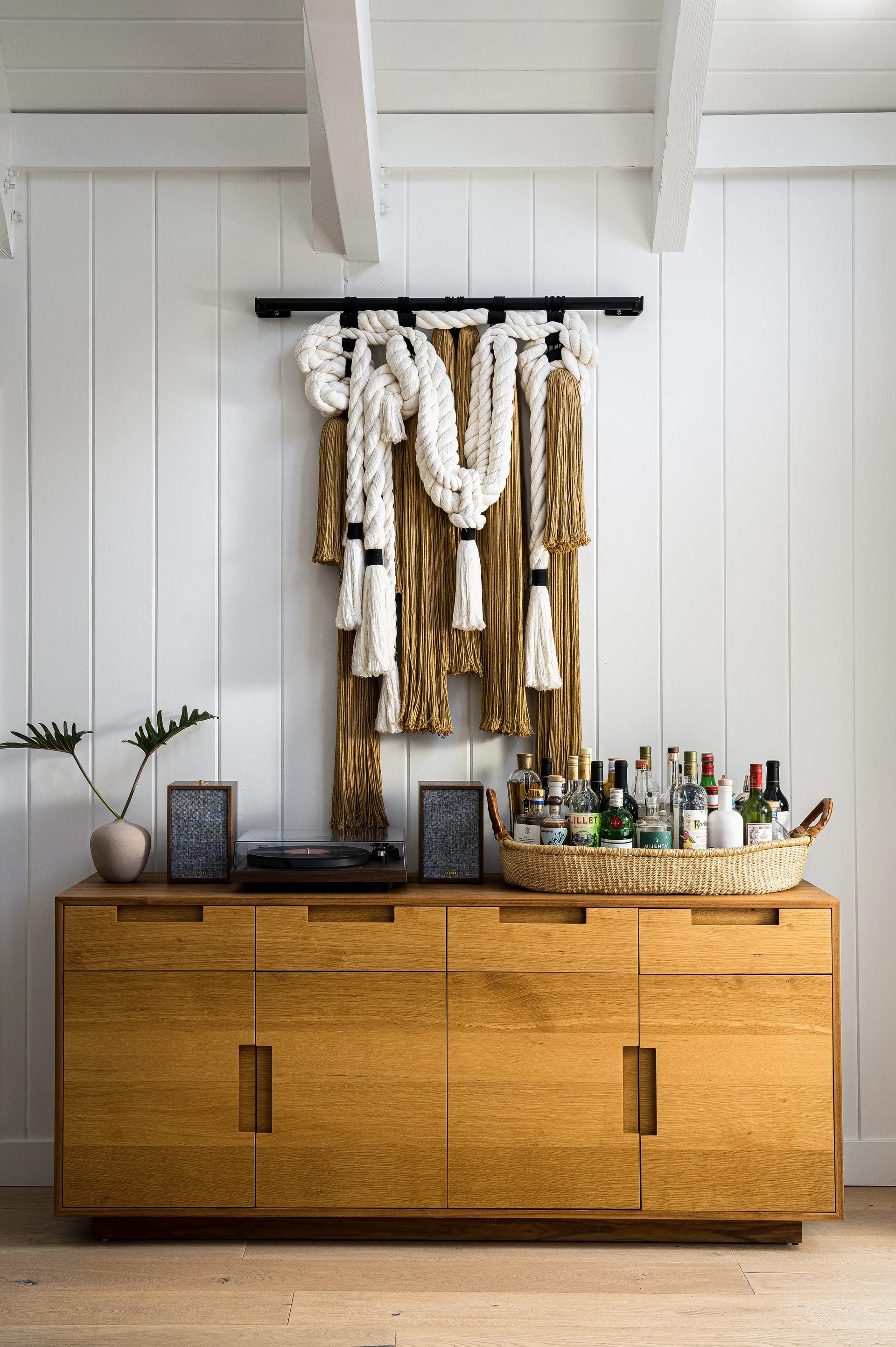 A wooden sideboard holds a turntable and a woven basket of liquor bottles beneath a large rope textile wall hanging on a white paneled wall.