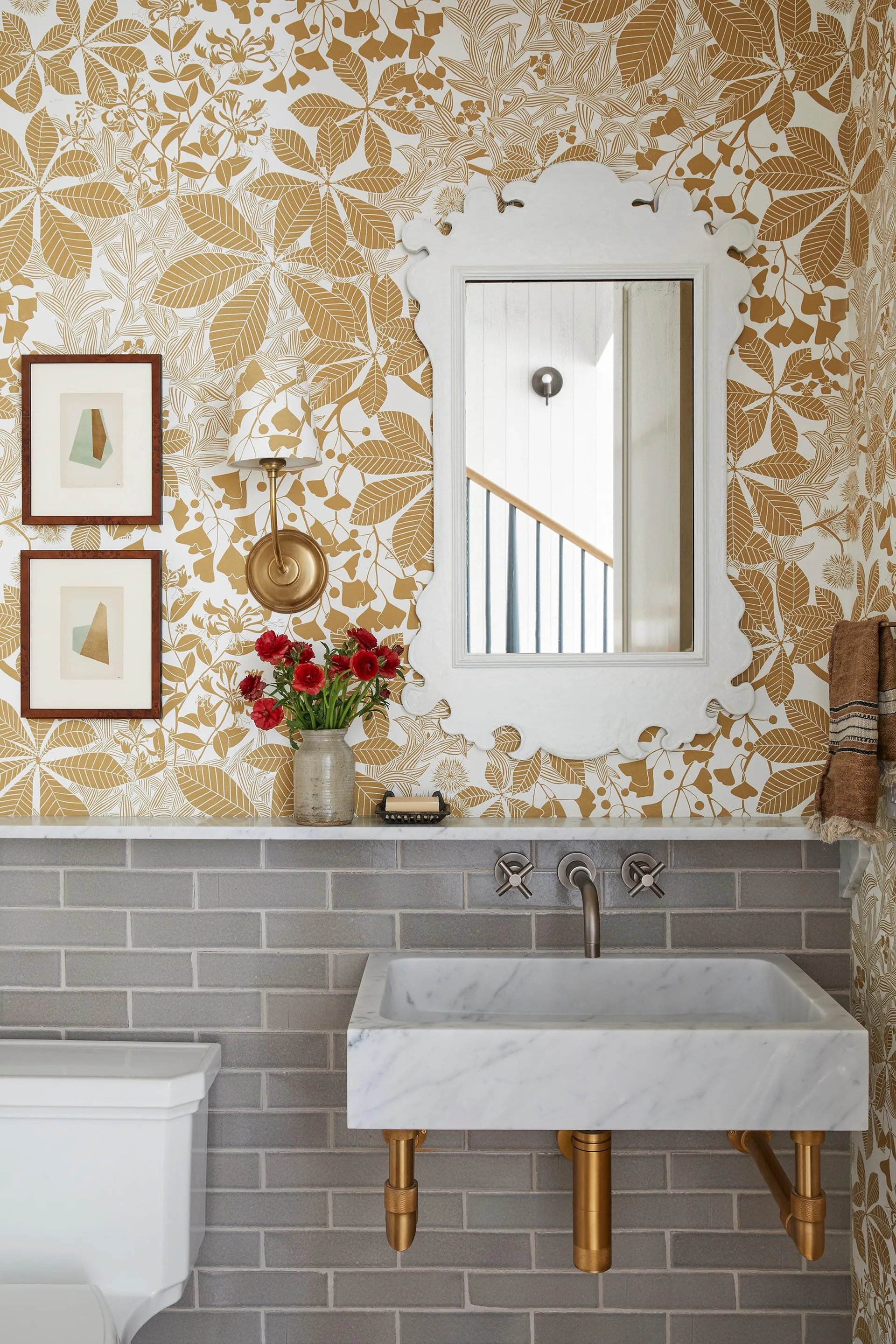 A powder room features gold and white leaf wallpaper, grey subway tiles, a wall-mounted marble sink with brass pipes, and an ornate white mirror.