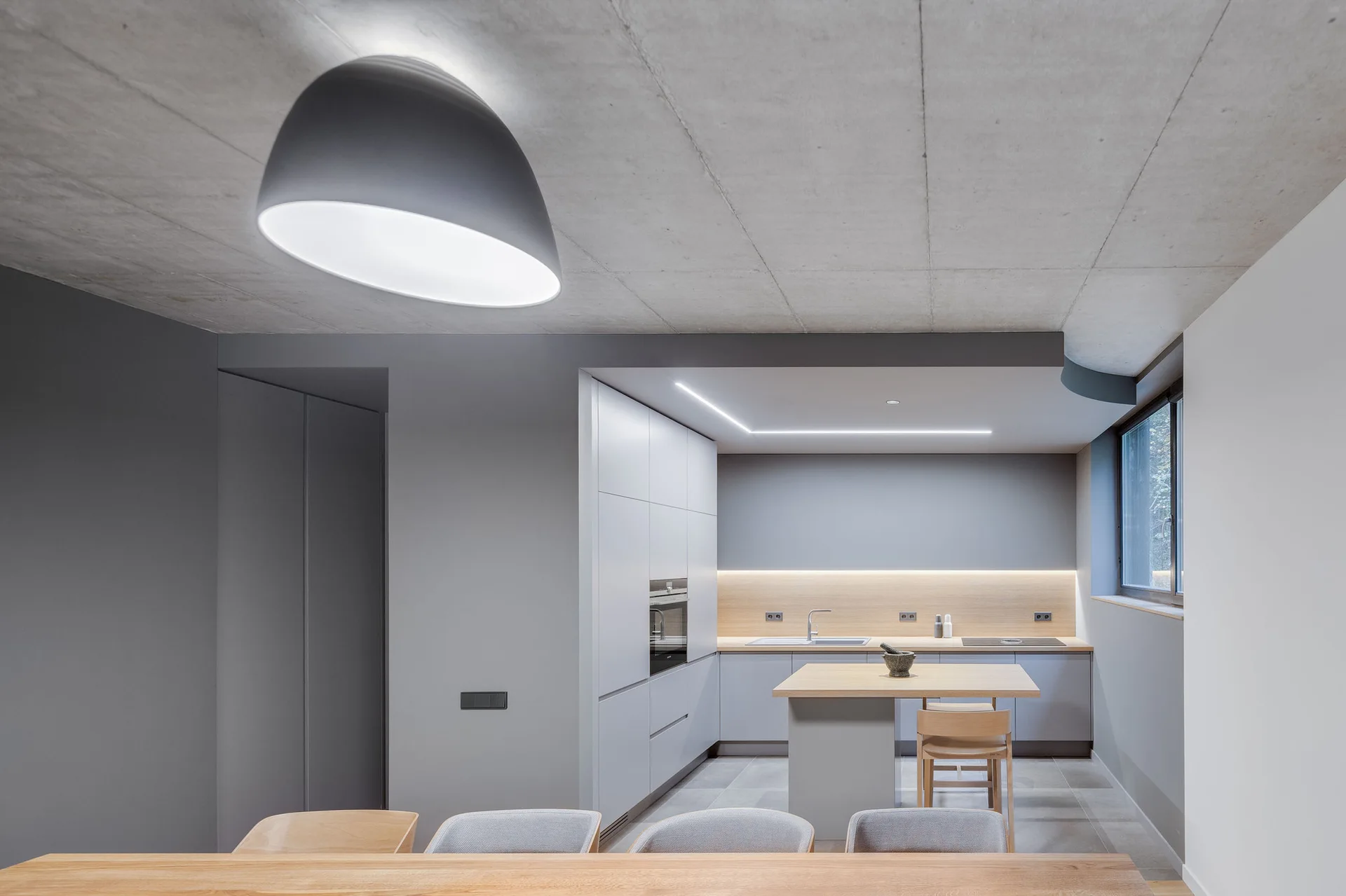 A minimalist kitchen with gray flat-panel cabinets, an exposed concrete ceiling, and a light wood-topped island with wooden bar stools.