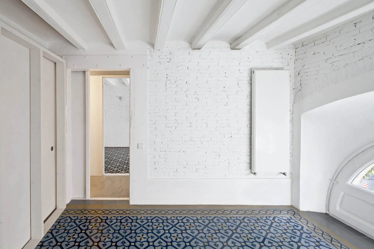 An empty room features white painted brick walls, exposed ceiling beams, and a blue and white patterned ceramic tile floor.