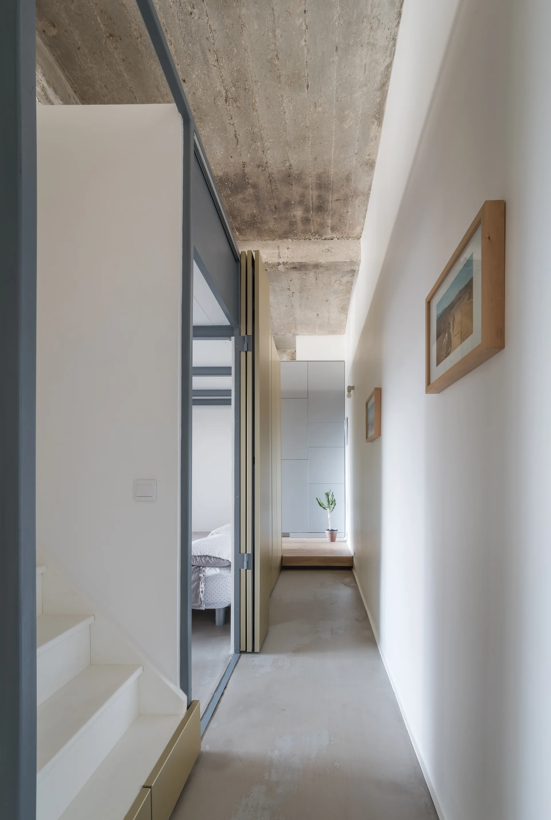 A narrow minimalist hallway features crisp white walls, a raw exposed concrete ceiling, a white staircase, and a folding partition door.