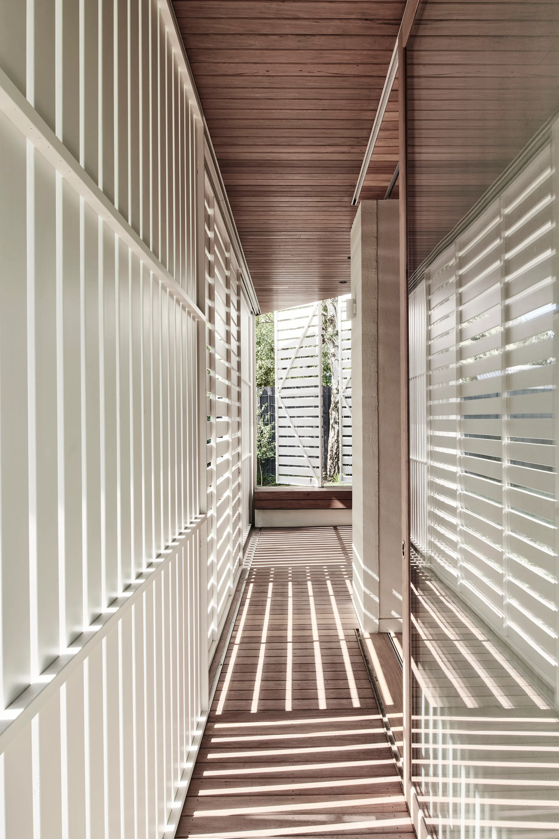 A narrow outdoor walkway features a warm wood-plank ceiling and deck, bordered by vertical white slats that cast linear shadows in the sunlight.