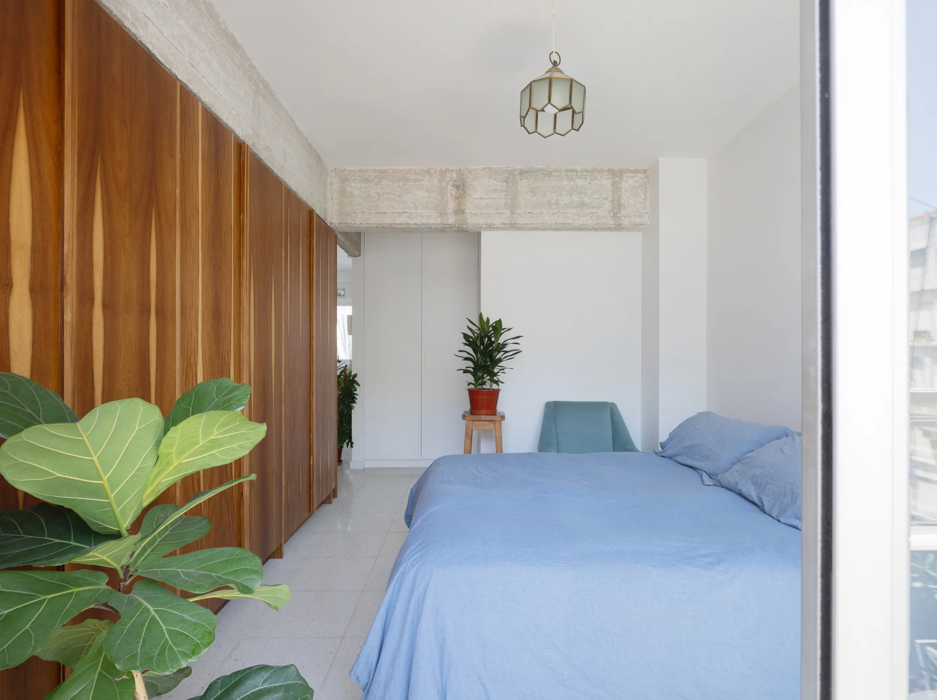 A minimalist bedroom featuring a bed with blue bedding, a large natural wood closet wall, and an exposed concrete beam overhead.
