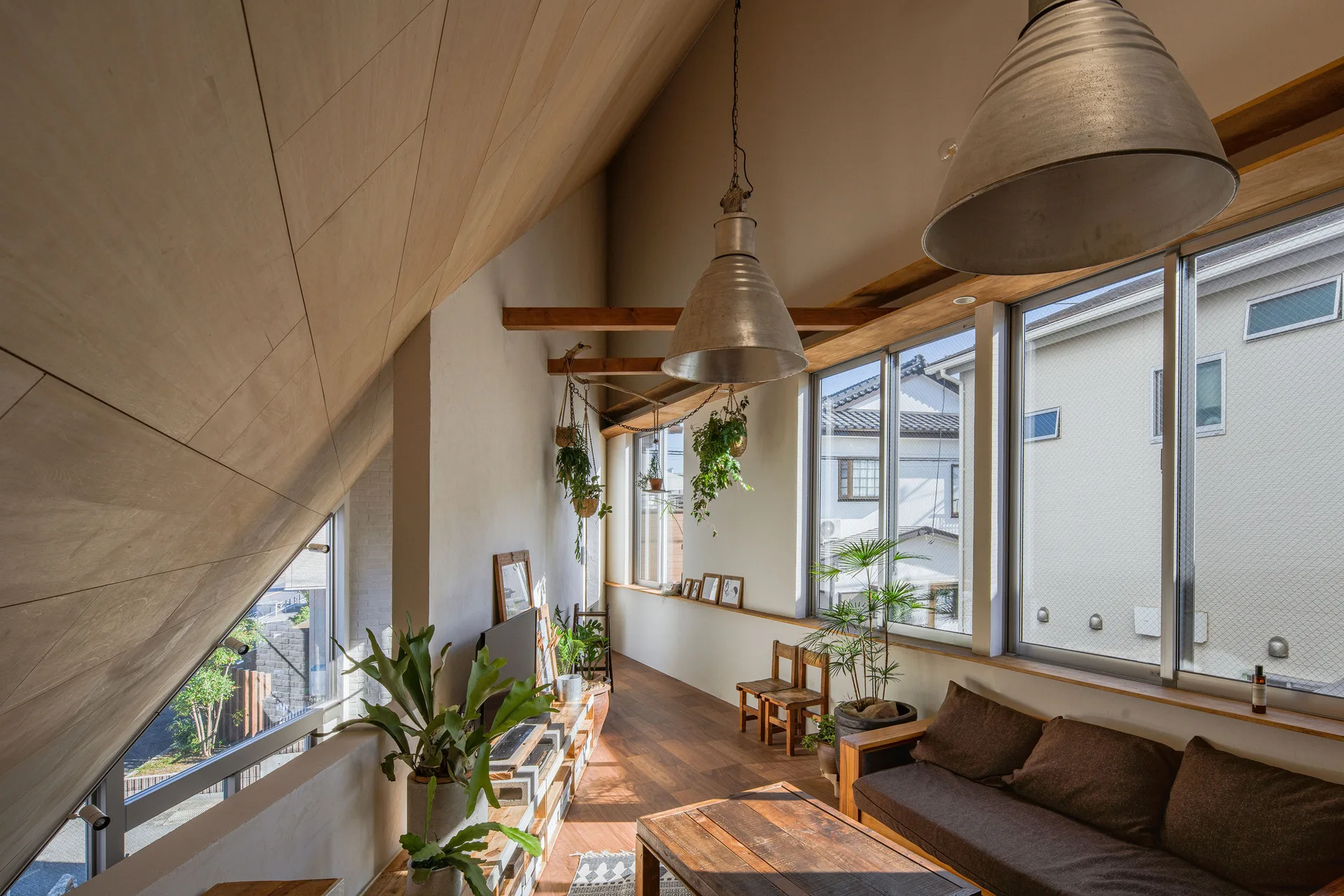 A lofted living room with a slanted wood panel ceiling, large windows overlooking neighborhood houses, and hanging green plants.