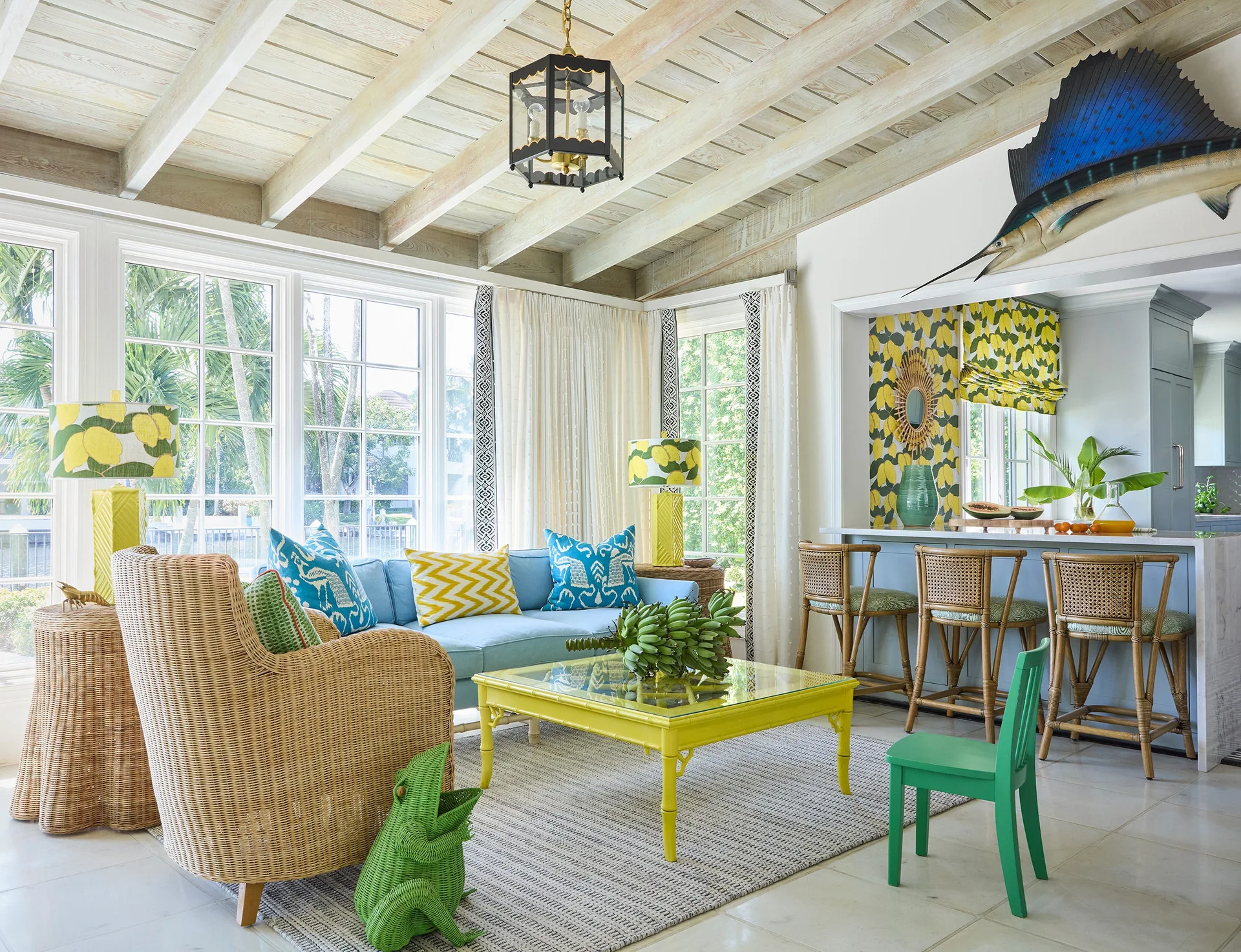 A coastal living room features a blue sofa, yellow coffee table, rattan armchair, and a kitchen counter with lemon-print wallpaper.