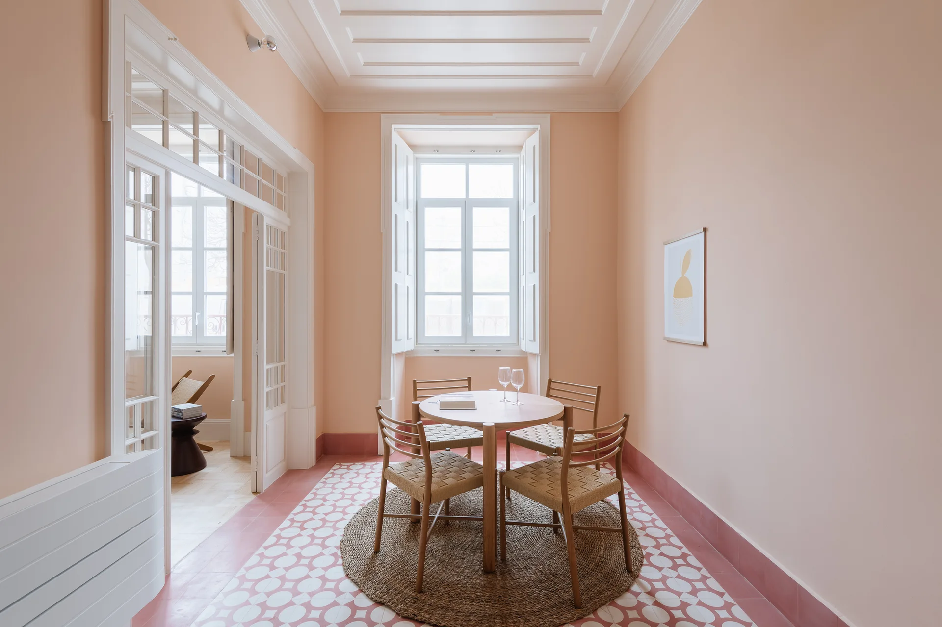 A dining space features peach walls, a round wooden table on a jute rug, and pink and white geometric floor tiles.