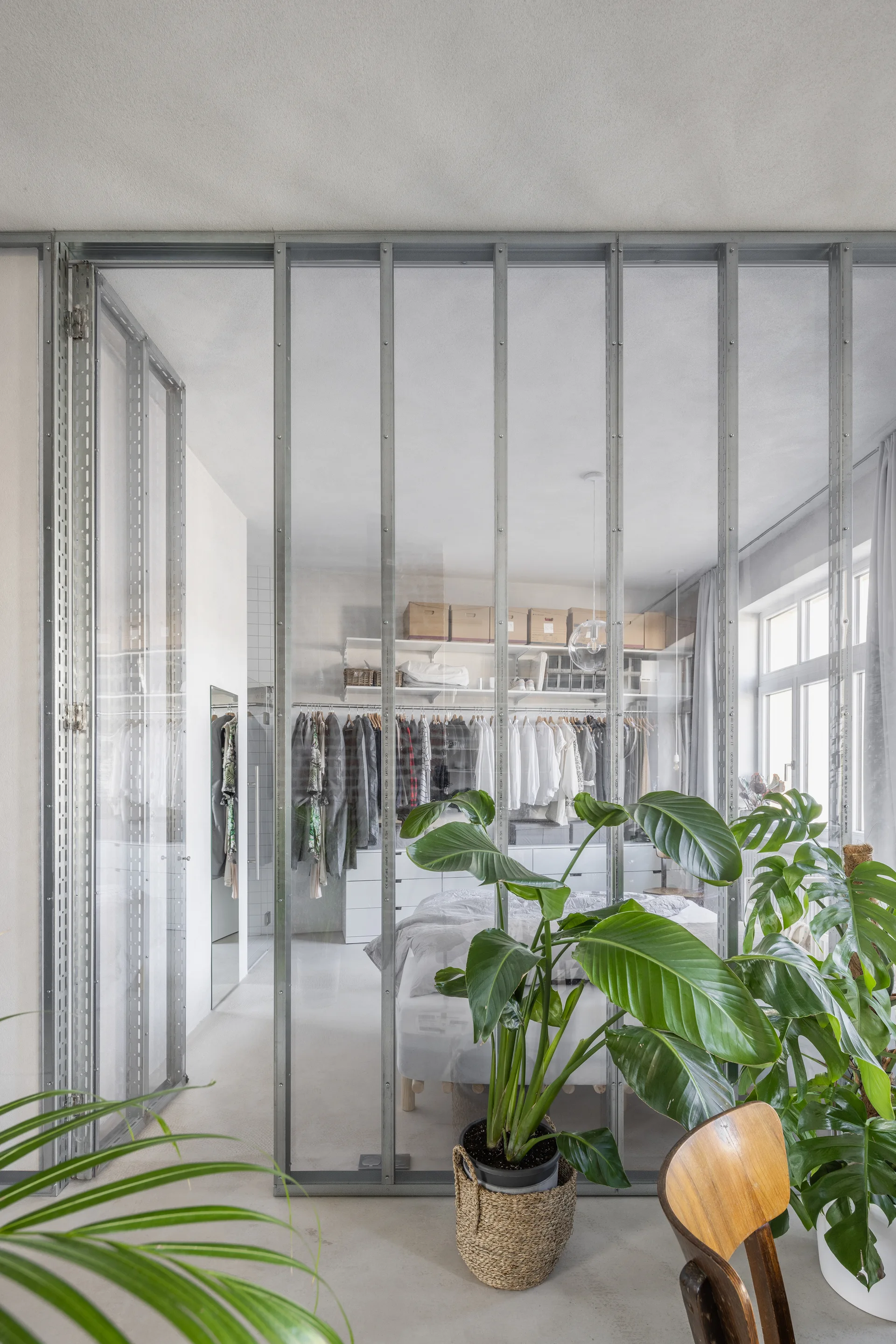 A tall metal-framed glass partition wall separates a room with green houseplants from a bedroom containing a bed and open clothing racks.