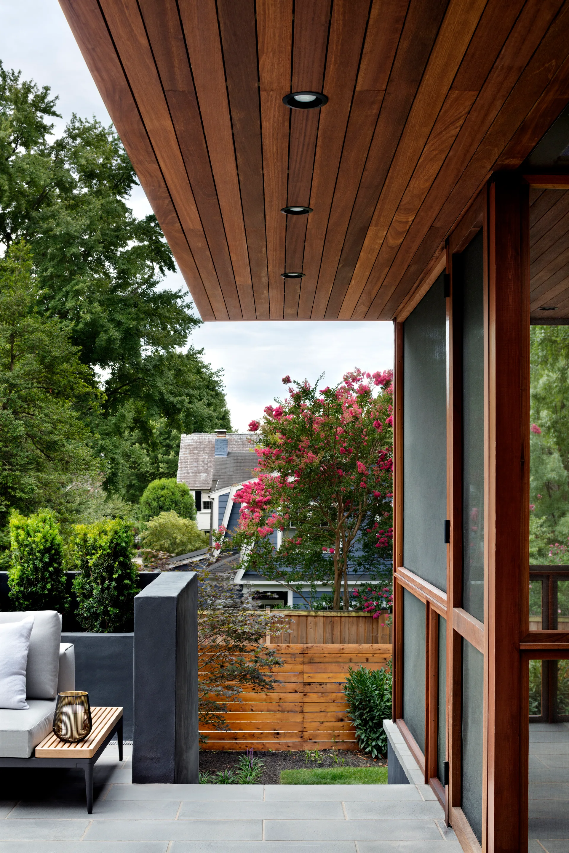 A covered stone patio features a grey cushioned sofa, a slatted wood side table, a built-in grey planter, and a wood plank ceiling.