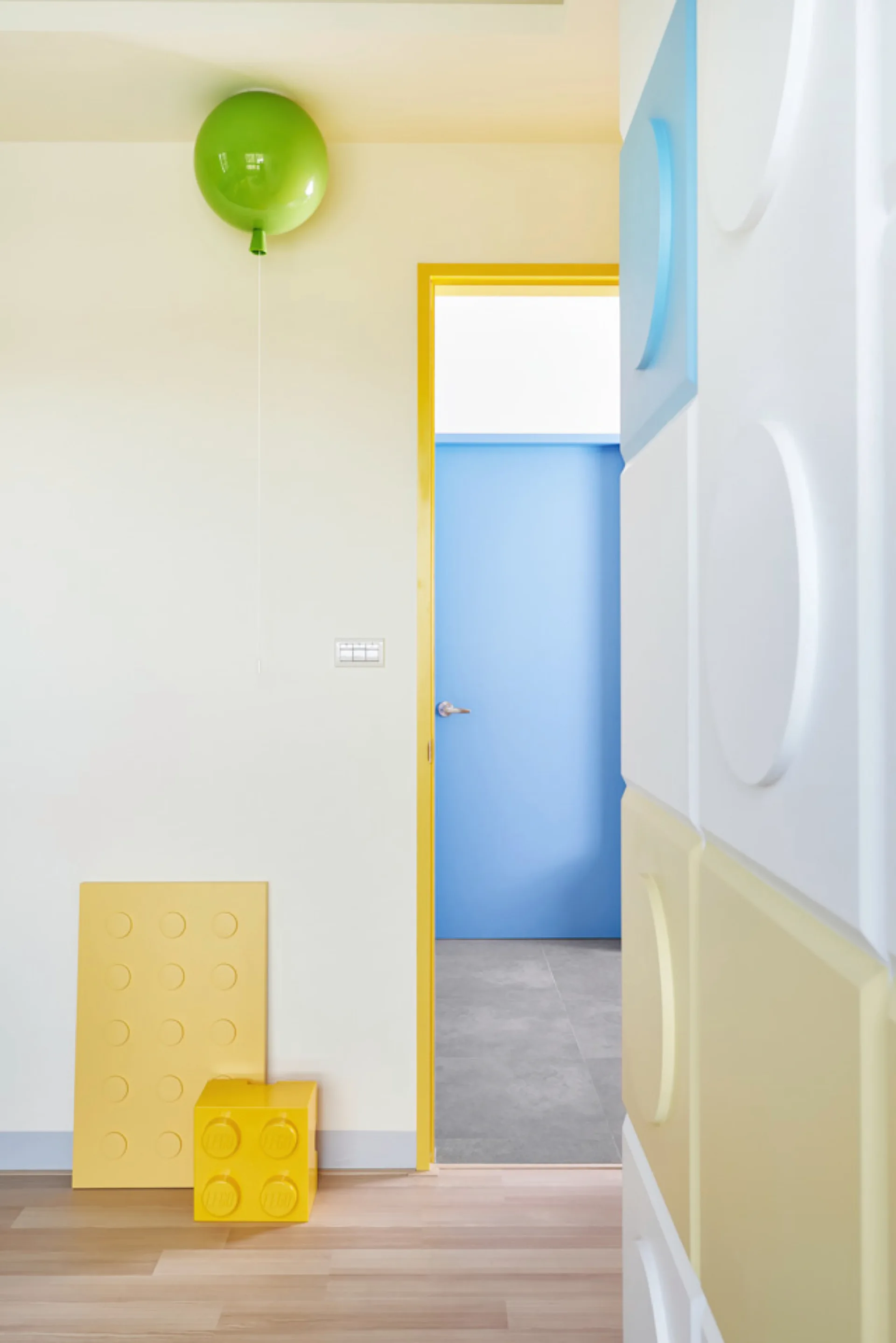 A playful playroom entry features a green balloon ceiling light, a bright yellow doorway leading to a blue door, and modular wall panels.