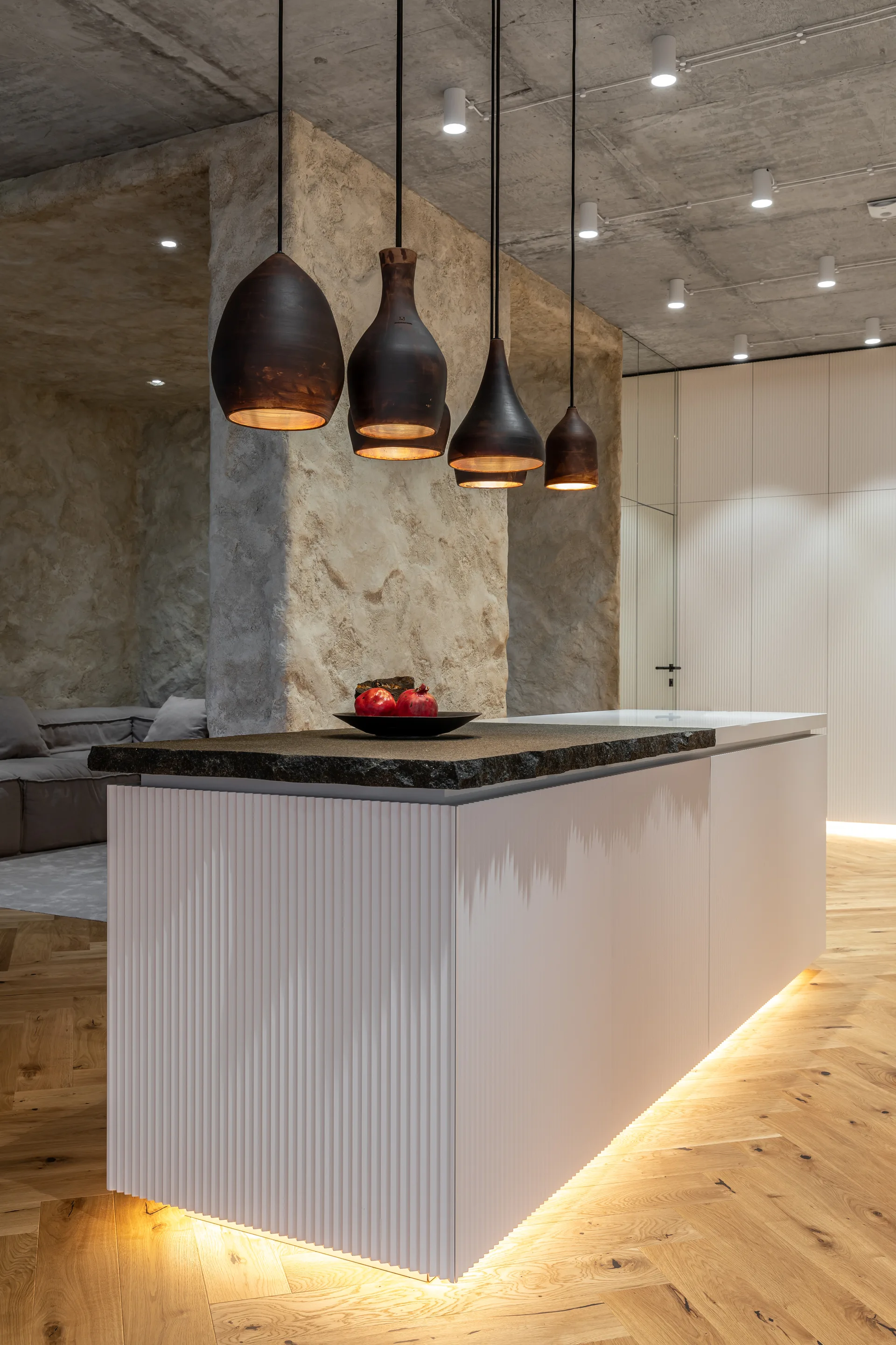 An off-white fluted kitchen island features a textured dark stone countertop beneath four hanging pendant lights and an exposed concrete ceiling.