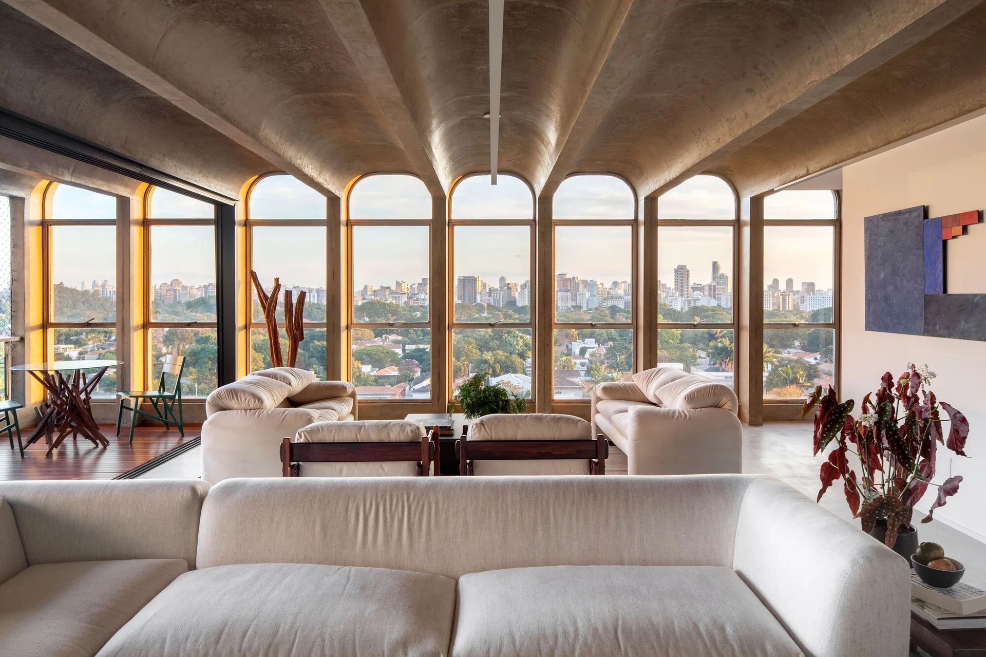 A spacious living room featuring a vaulted concrete ceiling, arched floor-to-ceiling windows with city views, and off-white sofas.