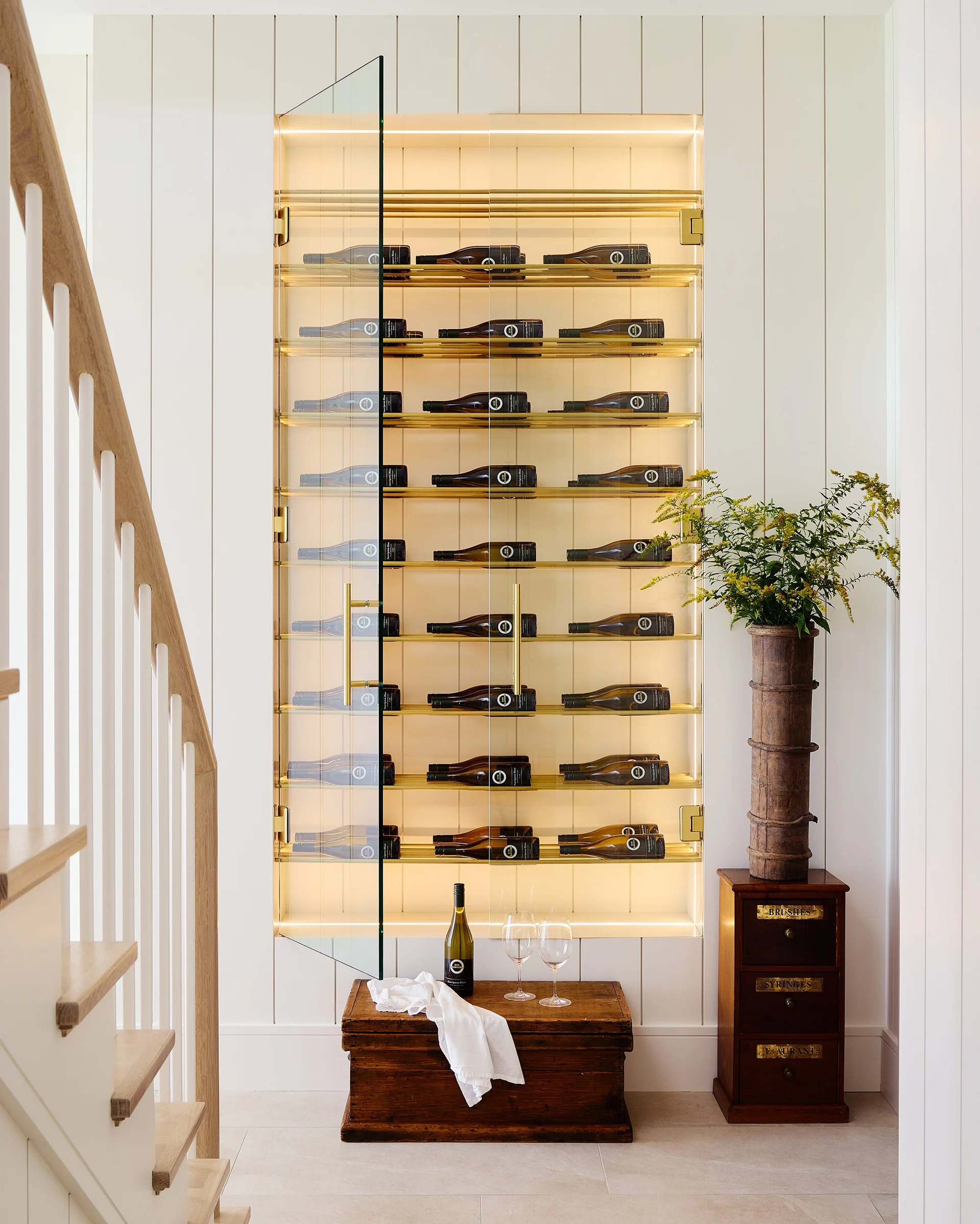 A backlit glass-front wine cabinet is built into a white vertical paneled wall next to a wooden staircase.