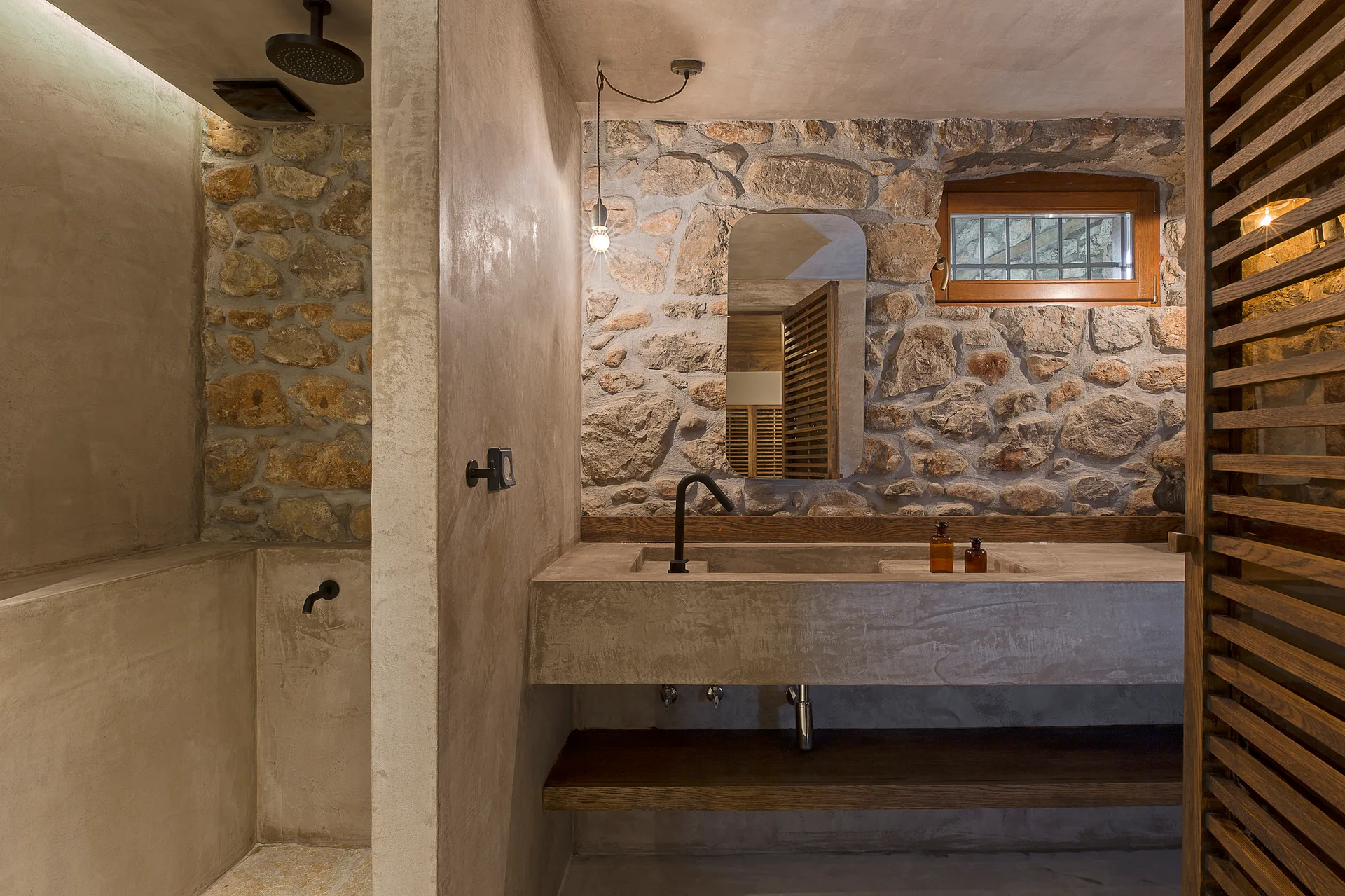 A rustic bathroom with a floating concrete vanity and matte black faucet set against a rough exposed stone wall next to a shower partition.