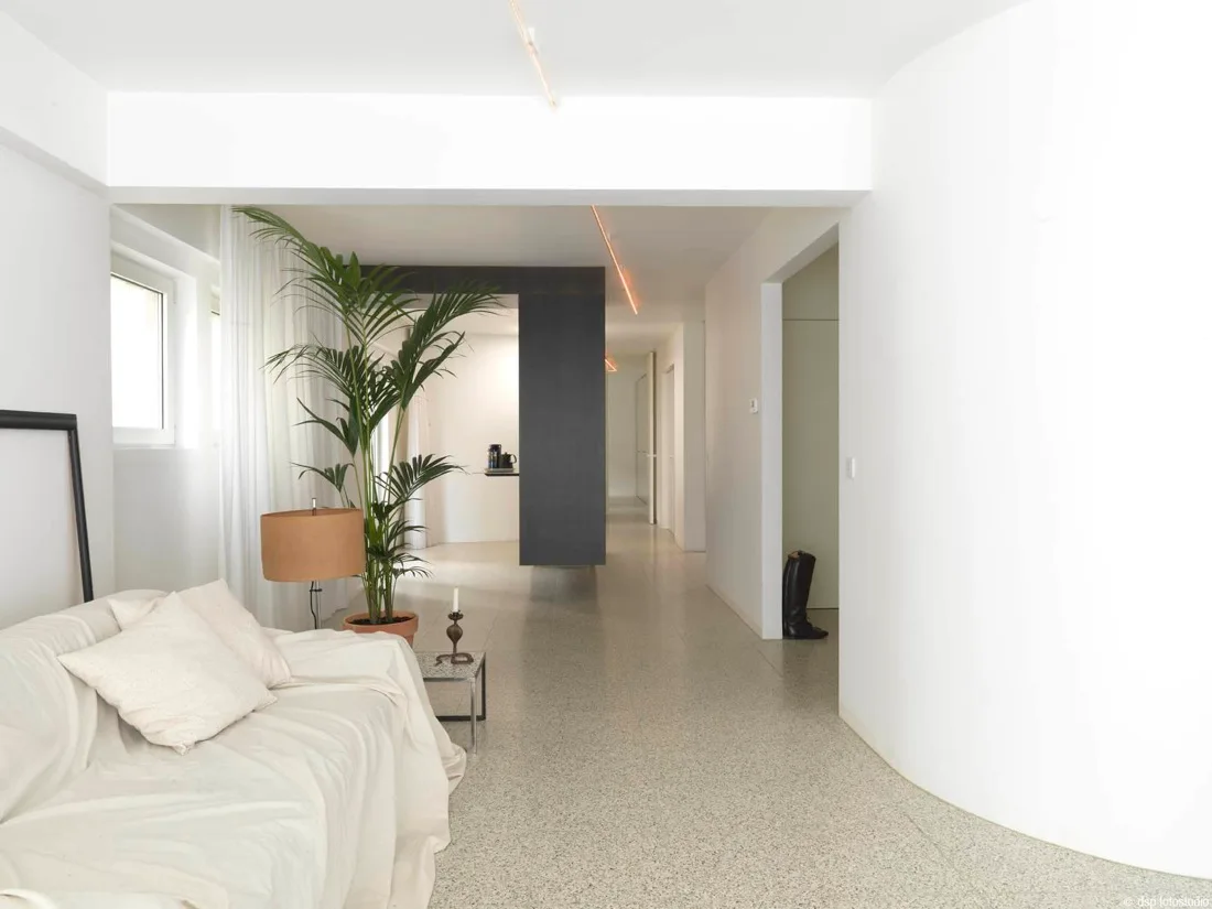 A minimalist living room with a white slipcovered sofa, a large potted plant, and light gray terrazzo flooring leading to a curved wall.