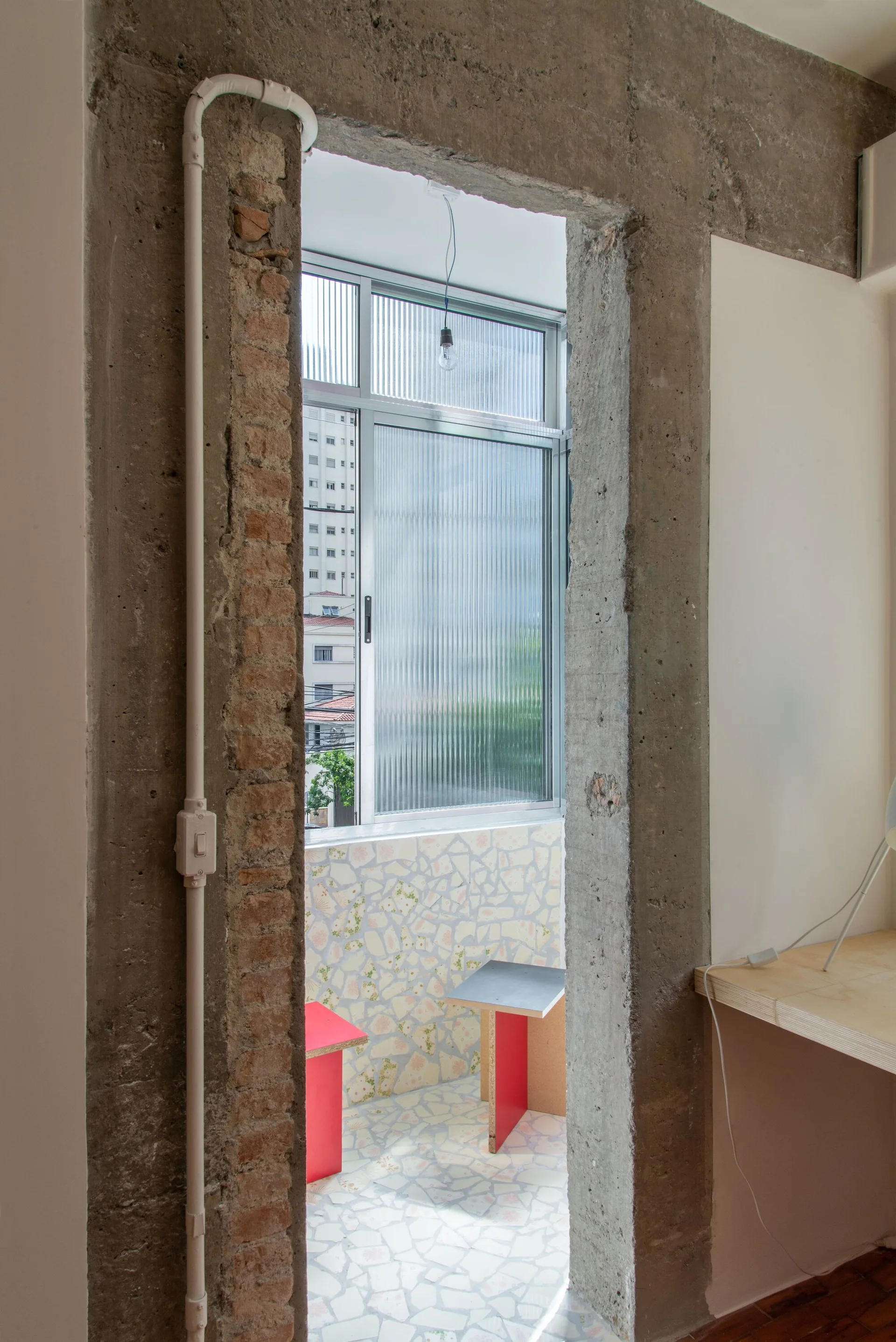 A view through an unfinished concrete doorway shows a bright balcony with a colorful mosaic tile floor, two stools, and ribbed glass windows.