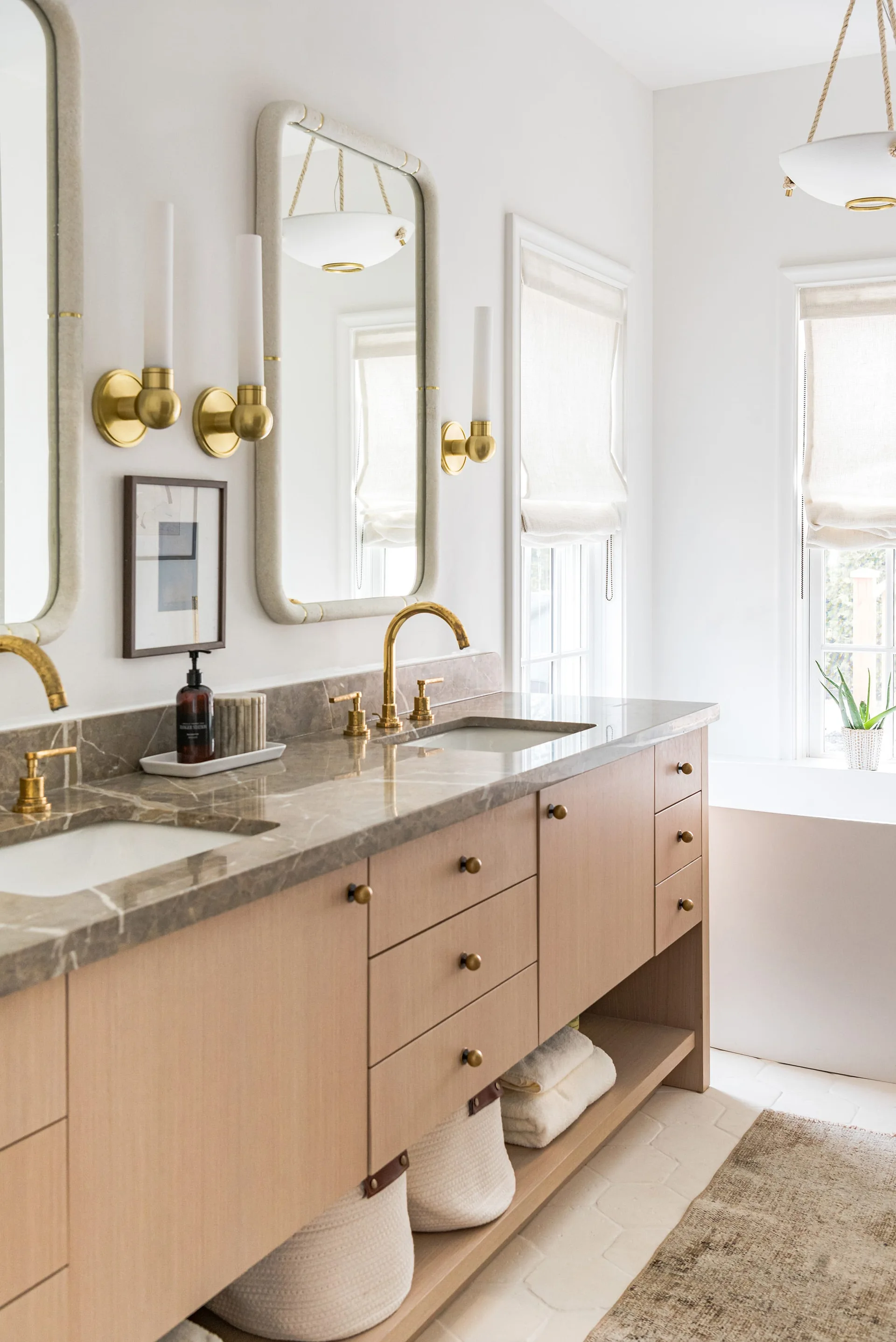 A light wood double vanity with a grey veined stone countertop, brass faucets, rounded rectangular mirrors, and brass sconces.