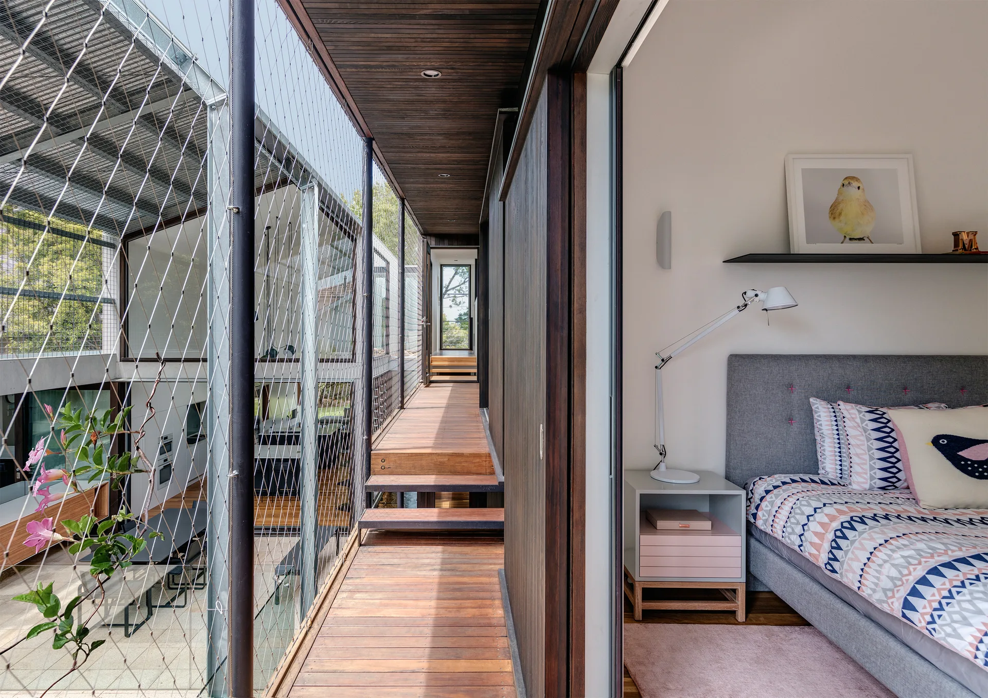 A bedroom with a grey upholstered bed and nightstand opens onto an elevated wooden walkway enclosed by a protective metal mesh safety screen.