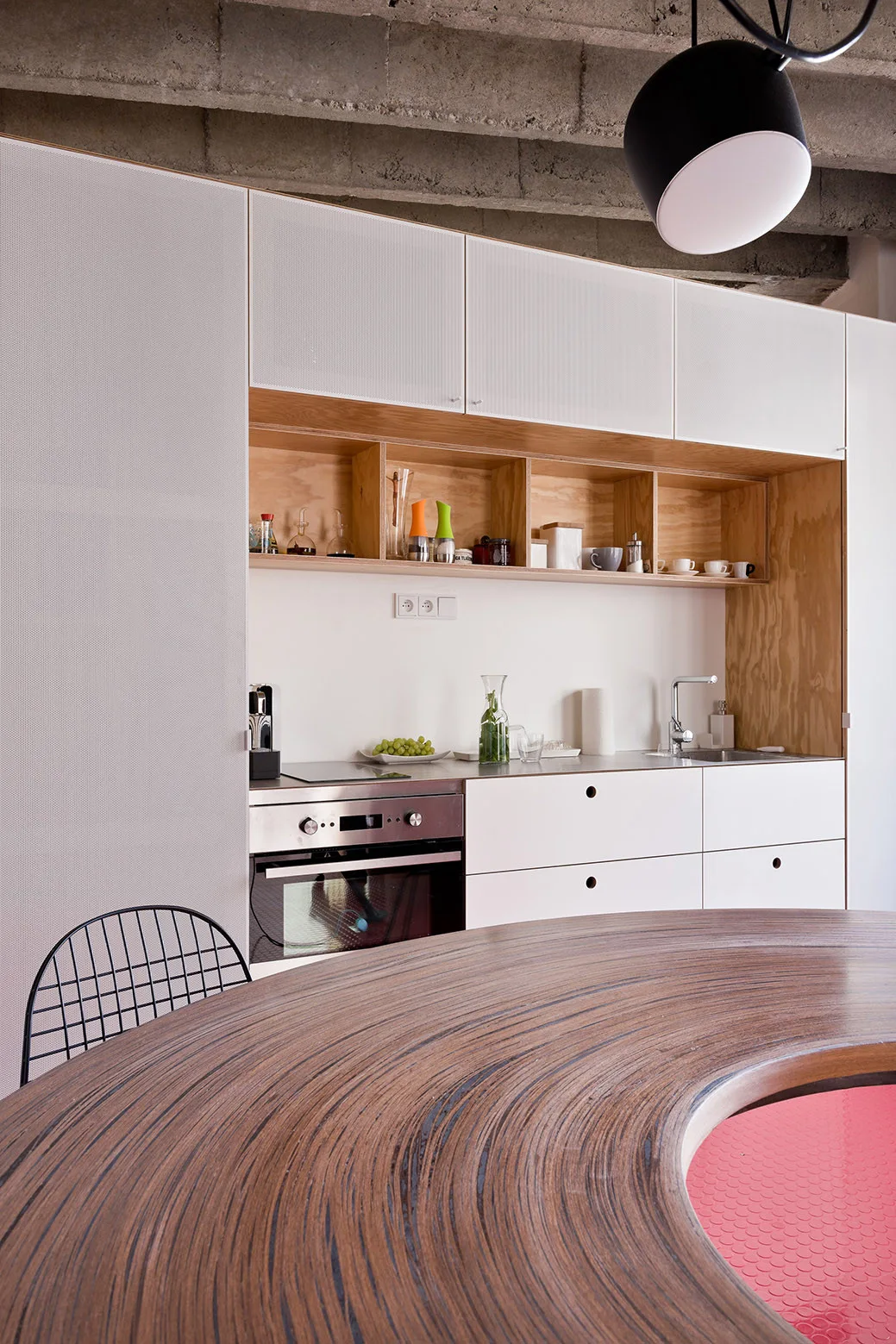 A minimalist kitchen features white cabinetry with an inset plywood shelf niche, concrete ceiling, and a curved wooden table in the foreground.