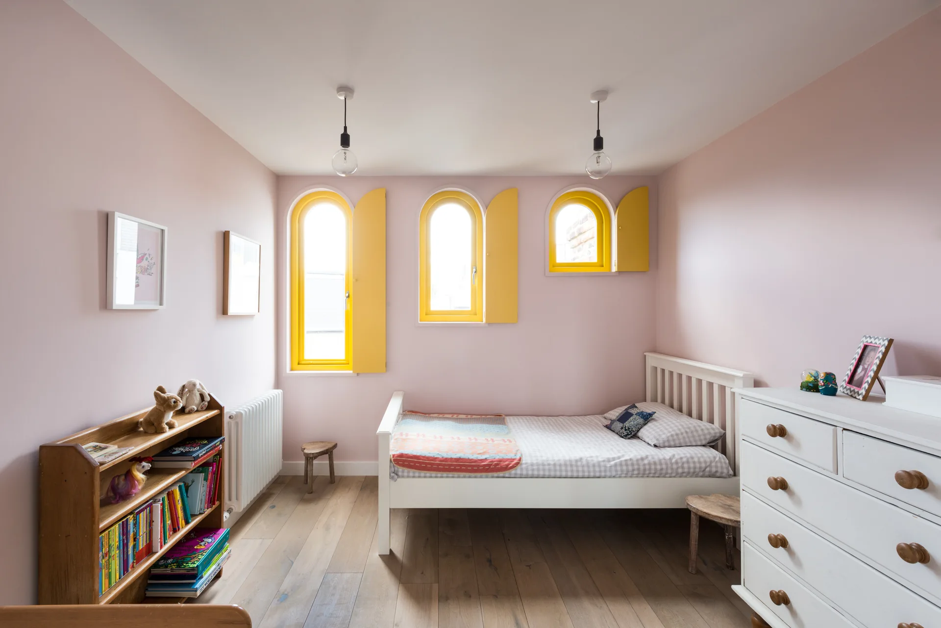 A children's bedroom features soft pink walls, a white wooden bed, a low bookshelf, and three arched windows with bright yellow shutters.