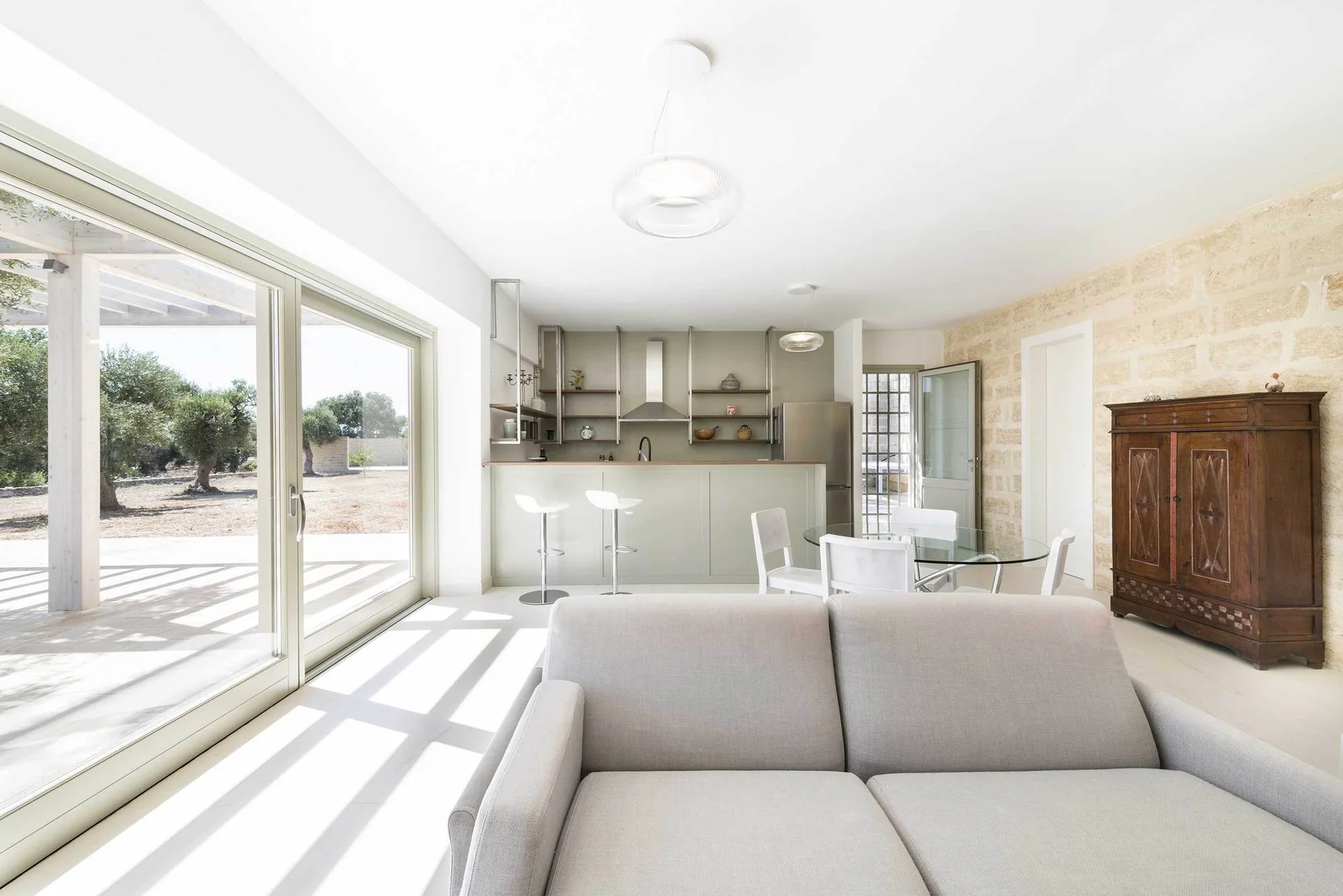 A minimalist open-plan living room with a light grey sofa, a sage green kitchen, and a textured stone wall.