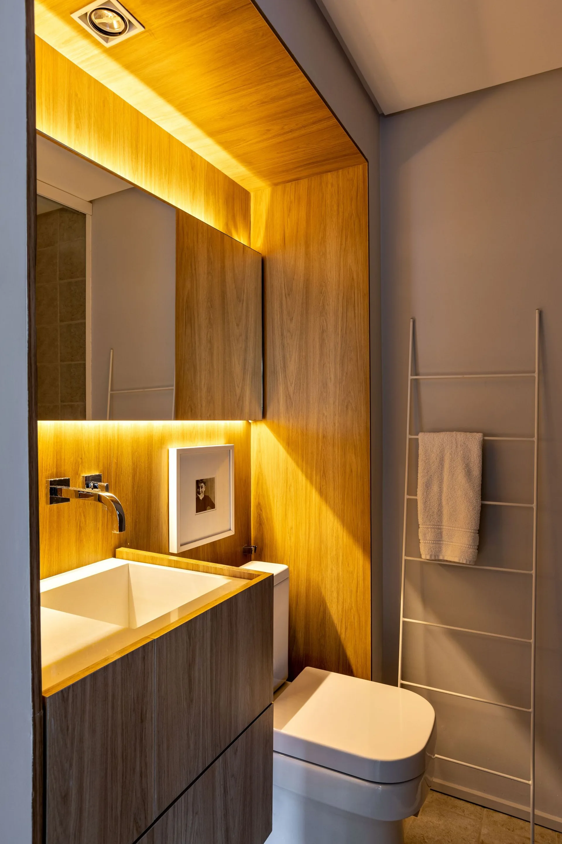 A modern powder room featuring a wood-veneered alcove with an integrated sink, a backlit mirror, a white toilet, and a white leaning towel ladder.