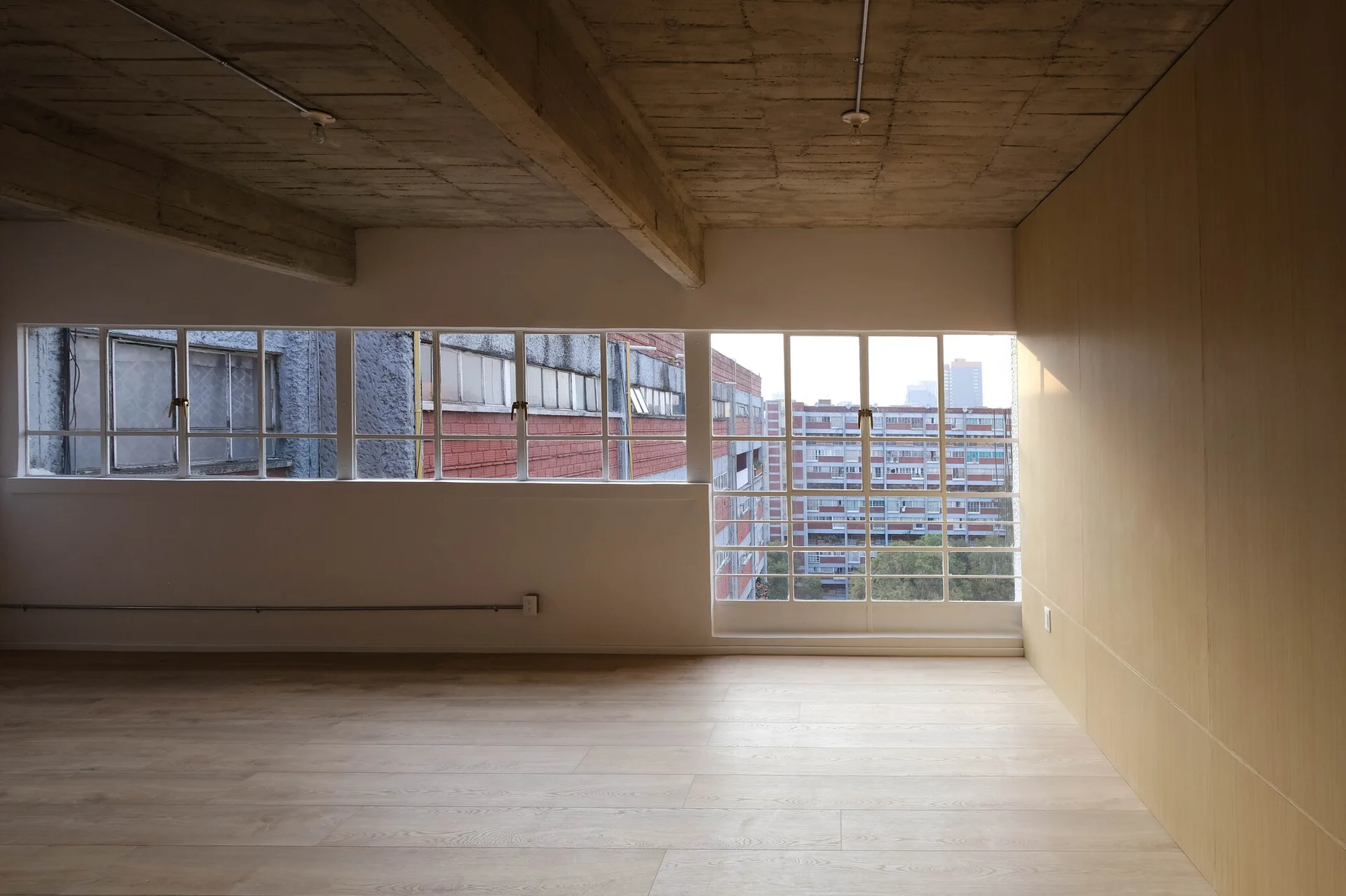 An empty minimalist apartment featuring a long horizontal grid window, light wood flooring, and an exposed raw concrete ceiling.