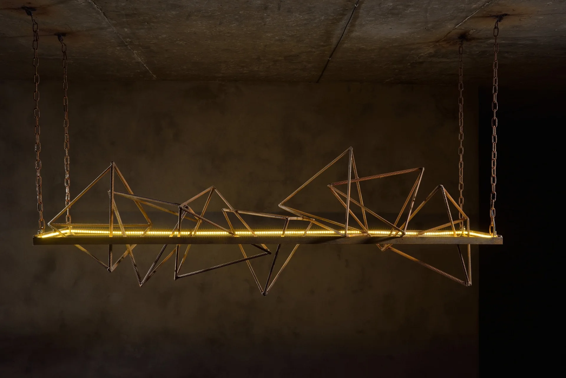 A sculptural geometric pendant light made of rusted rebar is suspended by chains from a concrete ceiling in front of a textured wall.