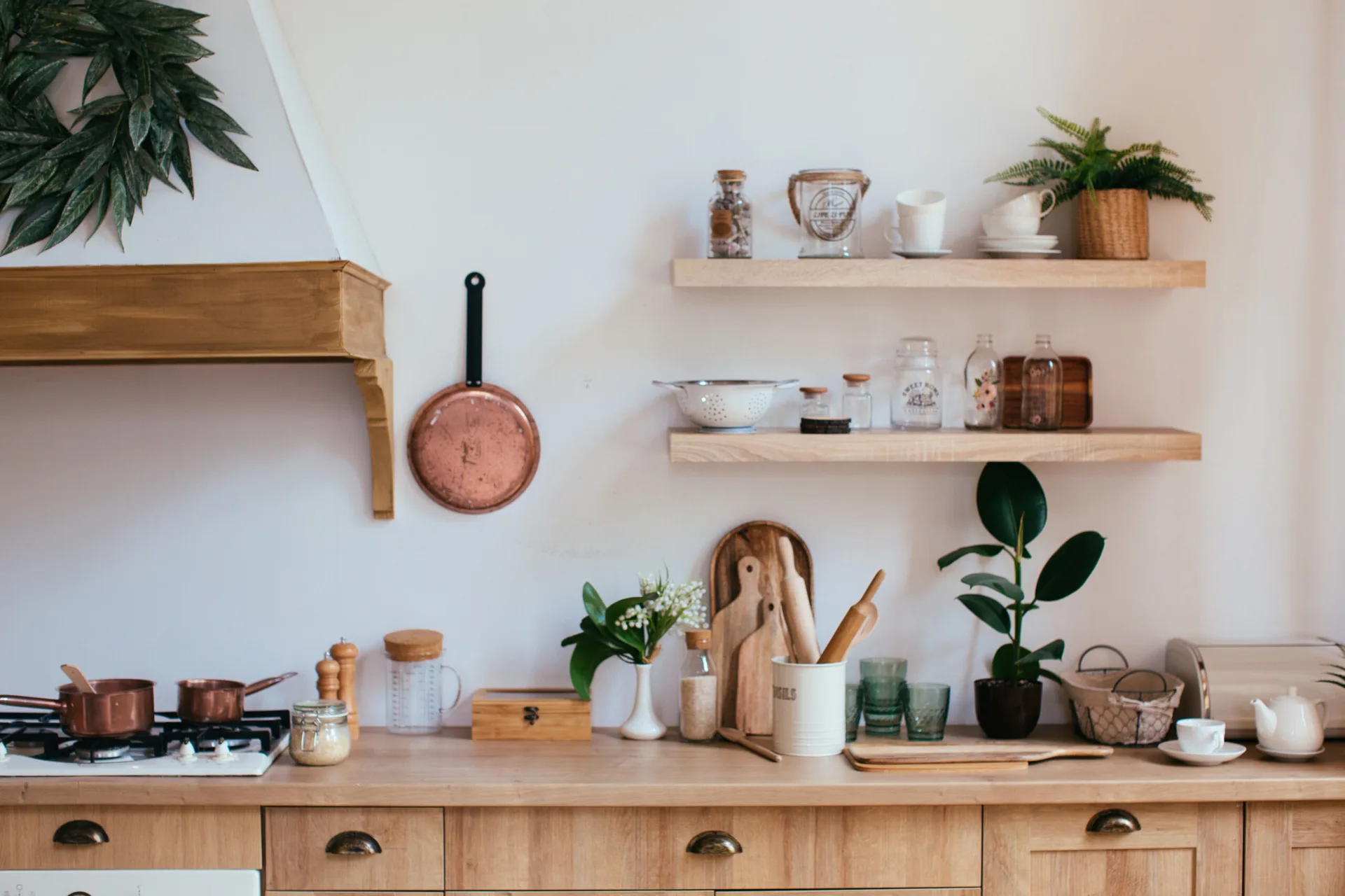 A rustic kitchen features light wood lower cabinets, two matching floating shelves with jars and ceramics, and copper pots on a gas stove.