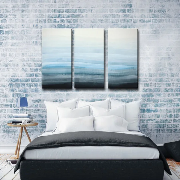 A bed with white pillows and a grey duvet sits in front of a whitewashed brick wall featuring a three-panel blue gradient art piece.