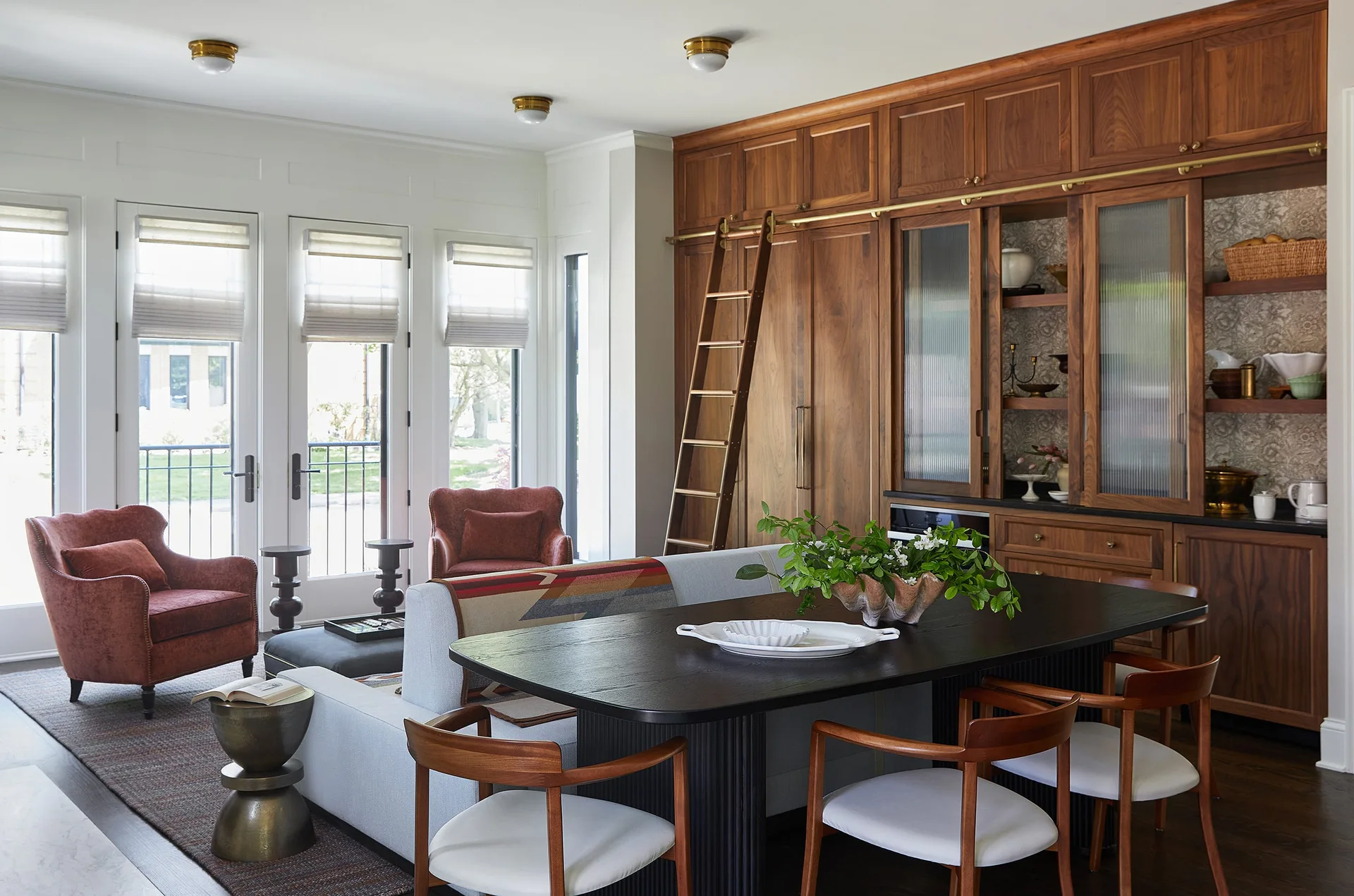 A transitional dining space featuring a black table, wooden chairs, a large walnut built-in wall unit with a library ladder, and adjacent armchairs.
